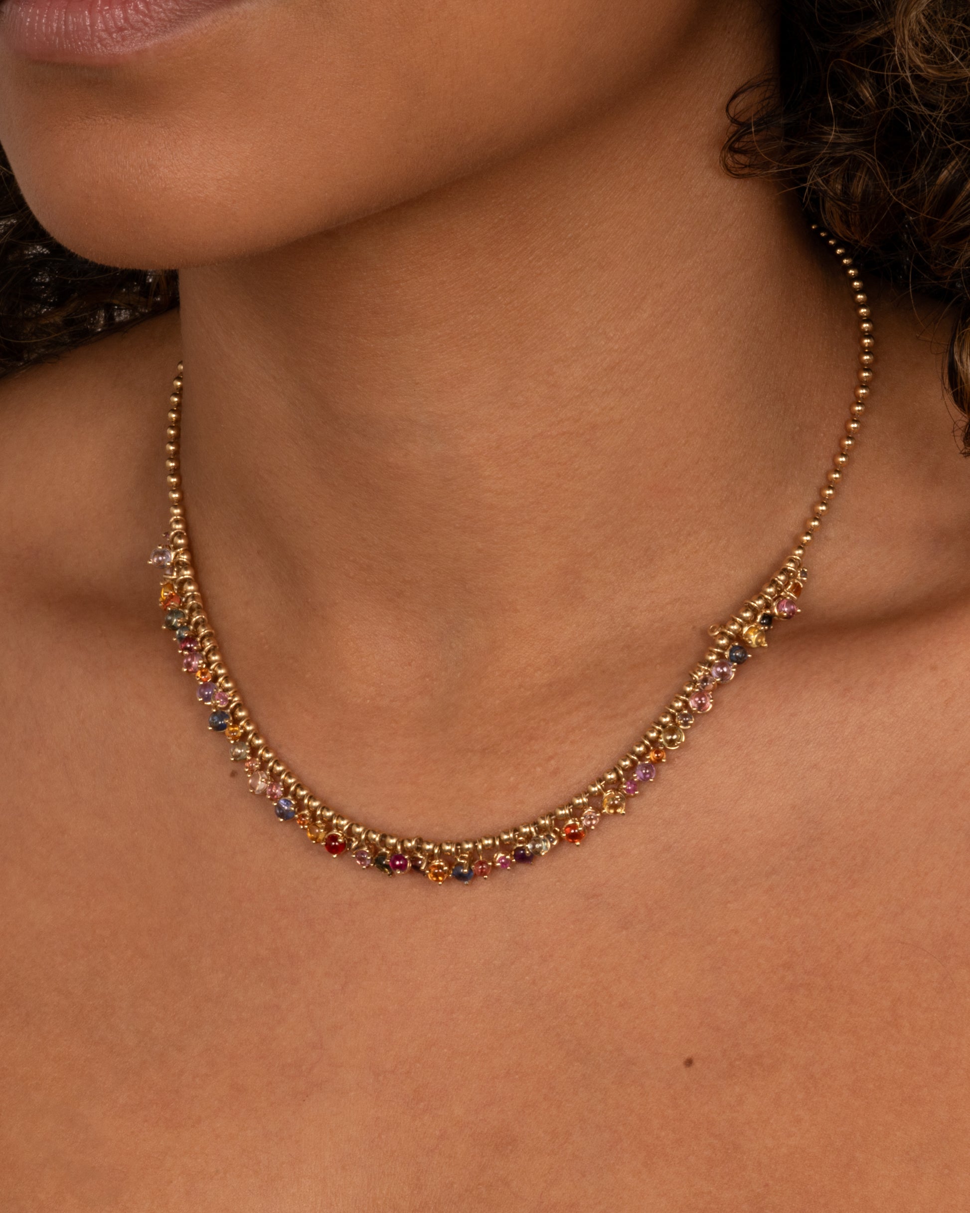 A close up of a yellow gold ball chain necklace with many multicolored sapphire dangles. Shown worn around the neck.