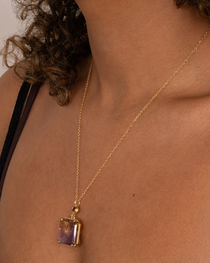 A yellow gold pendant necklace with an ametrine sunset carving with a gold sun face on its reverse. A round amethyst hovers just above this double sided pendant. Shown a person.