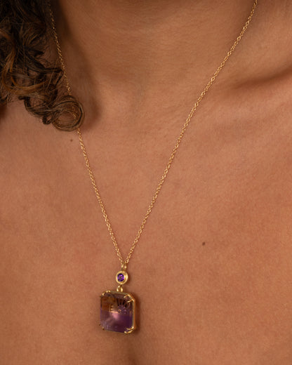 A yellow gold pendant necklace with an ametrine sunset carving with a gold sun face on its reverse. A round amethyst hovers just above this double sided pendant. Shown on a person.