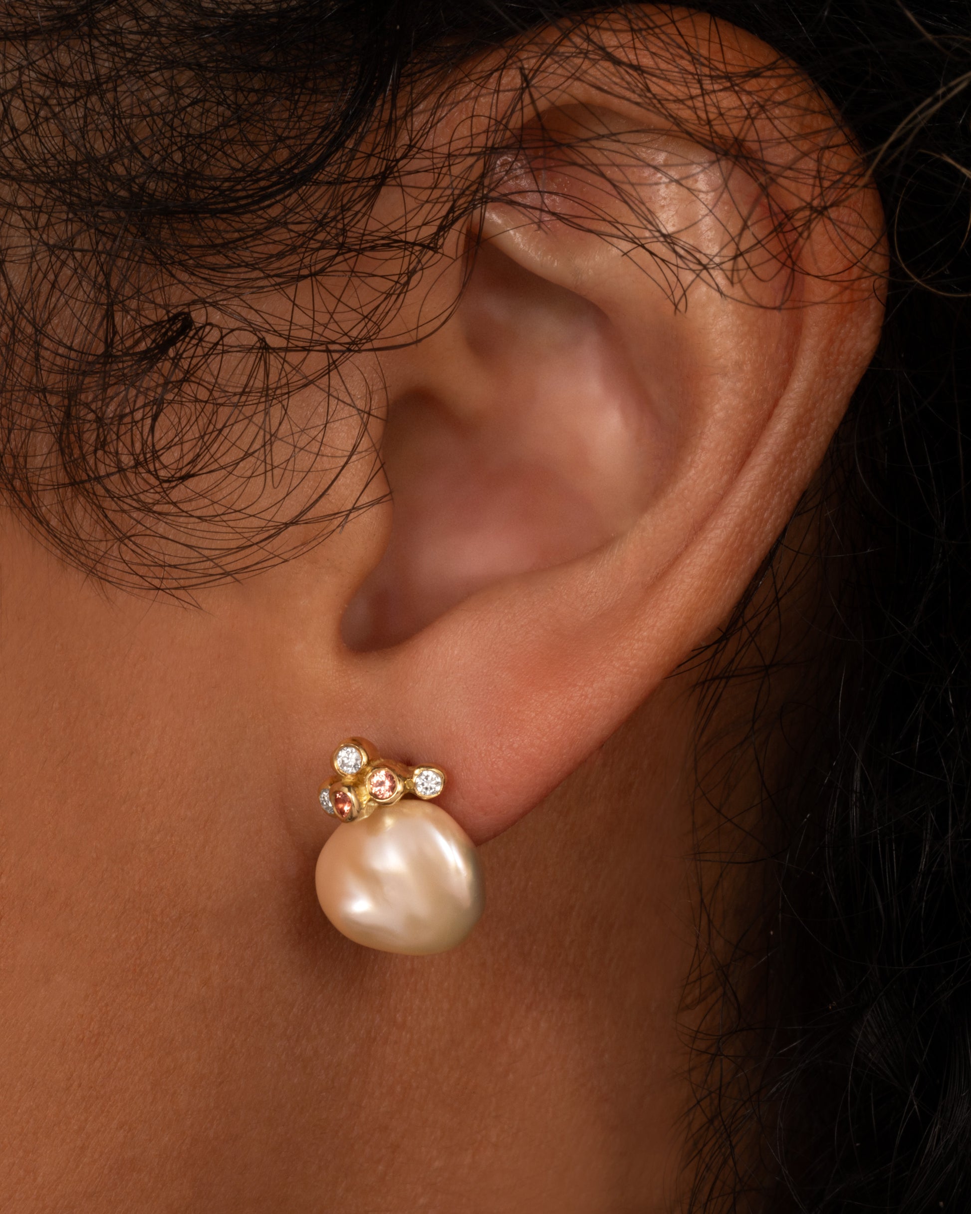 A yellow gold stud earring with a diamond and orange sapphire cluster atop a pink freeform Keshi pearl, shown on a person.