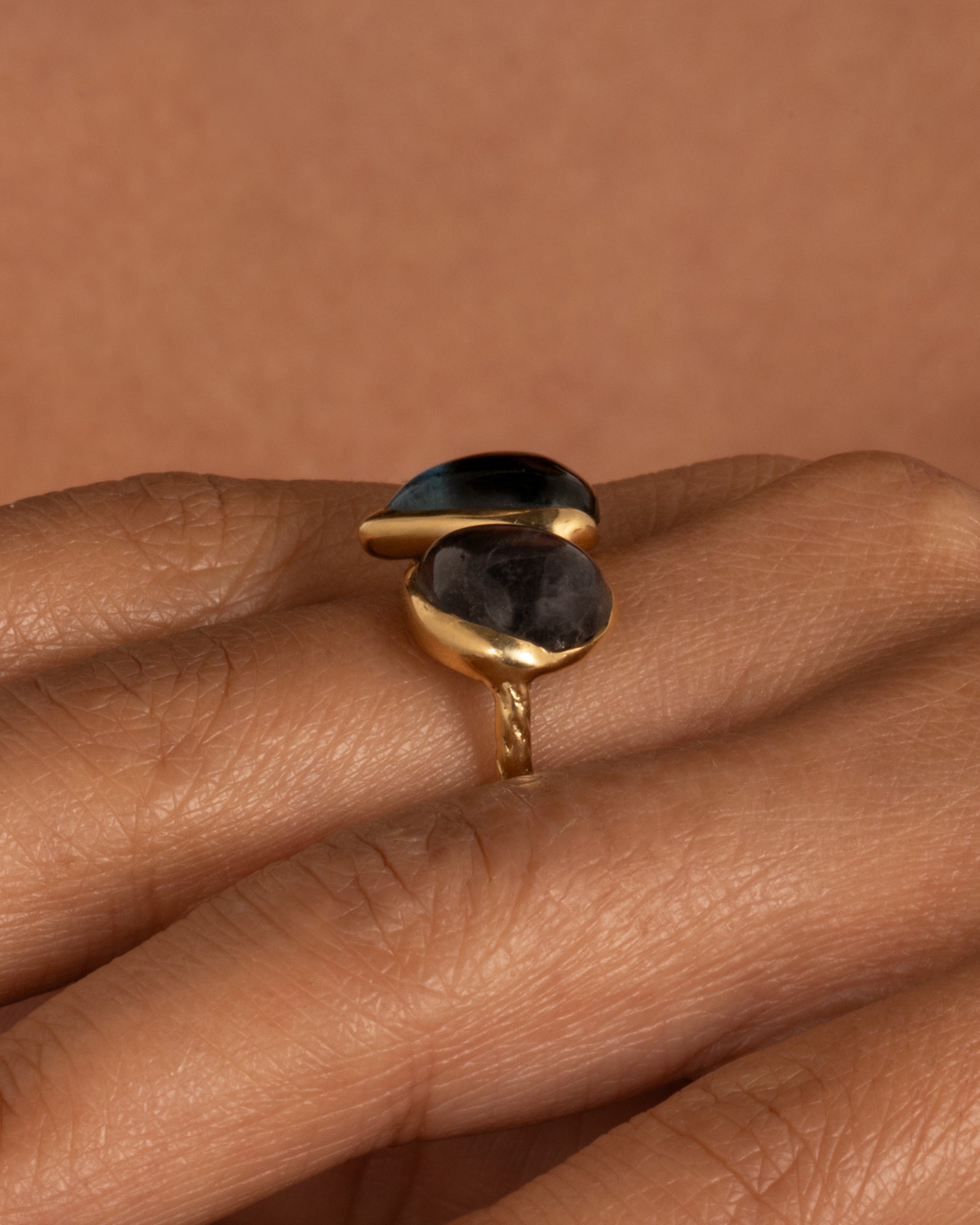 A yellow gold ring with a pair of blue green tourmalines and a textured hand- carved band, shown from the side on a hand.