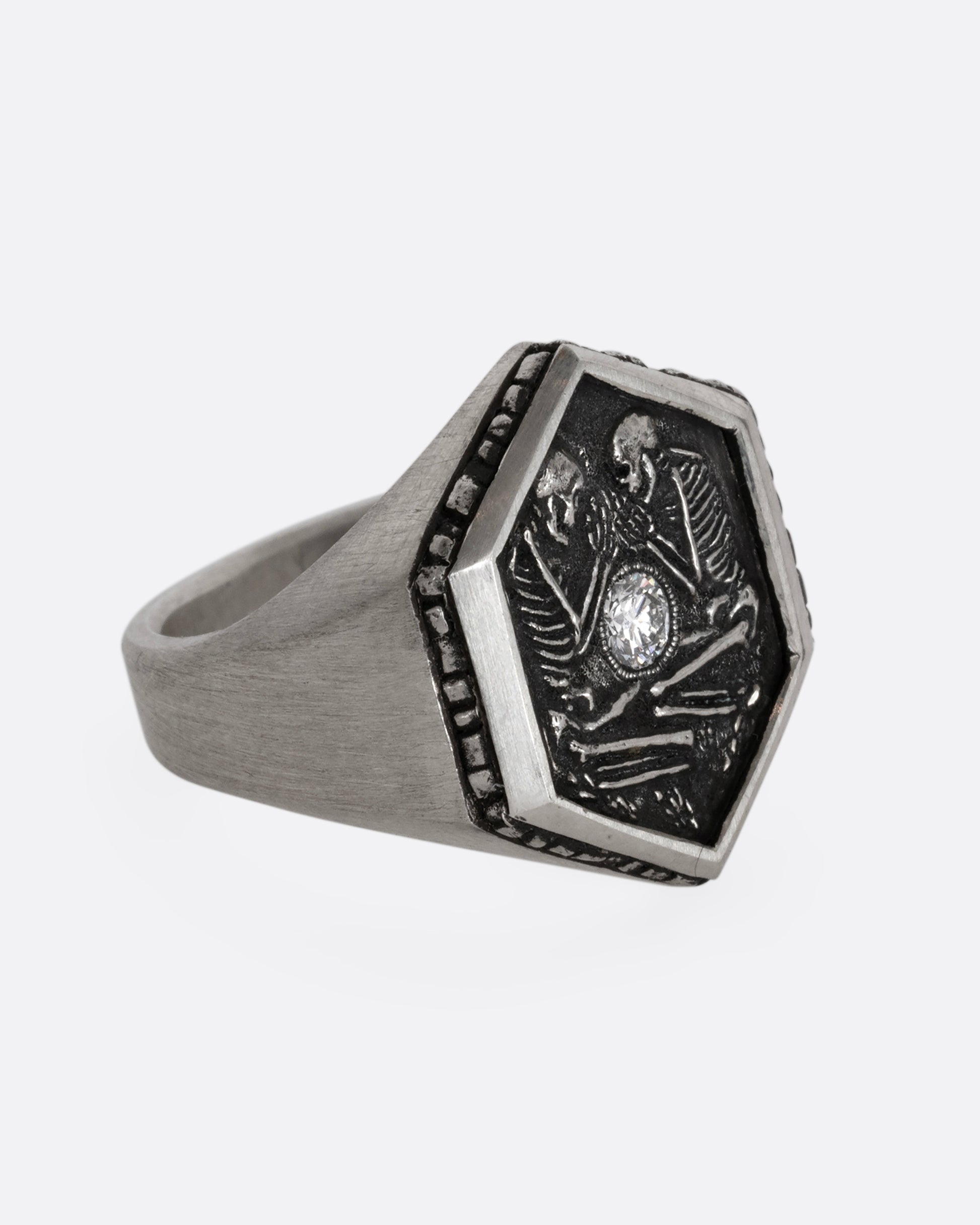 Sterling silver signet ring with two skeletons and a diamond at the center. Shown from the side.