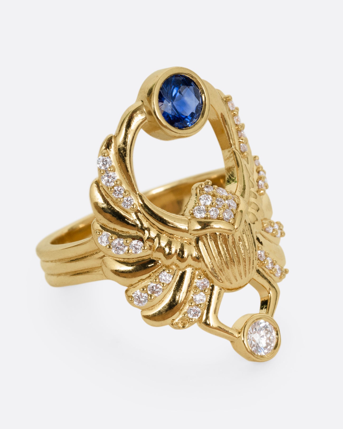 A yellow gold scarab ring with accents of white diamonds and a single blue sapphire between its fanned wings. Shown from the side.