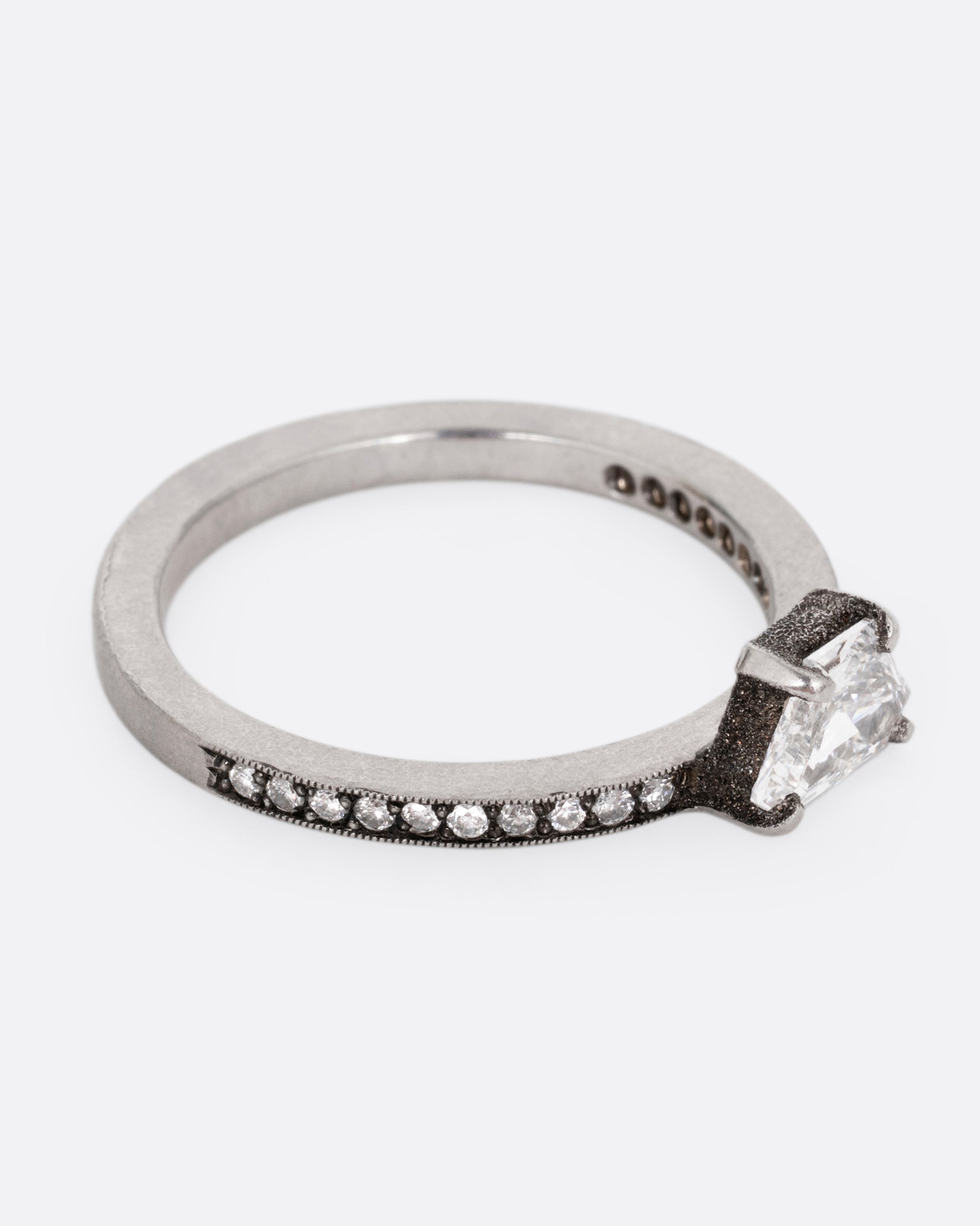 A platinum band with pave diamonds all the way around and a solitaire trapeze diamond. Shown from the side.