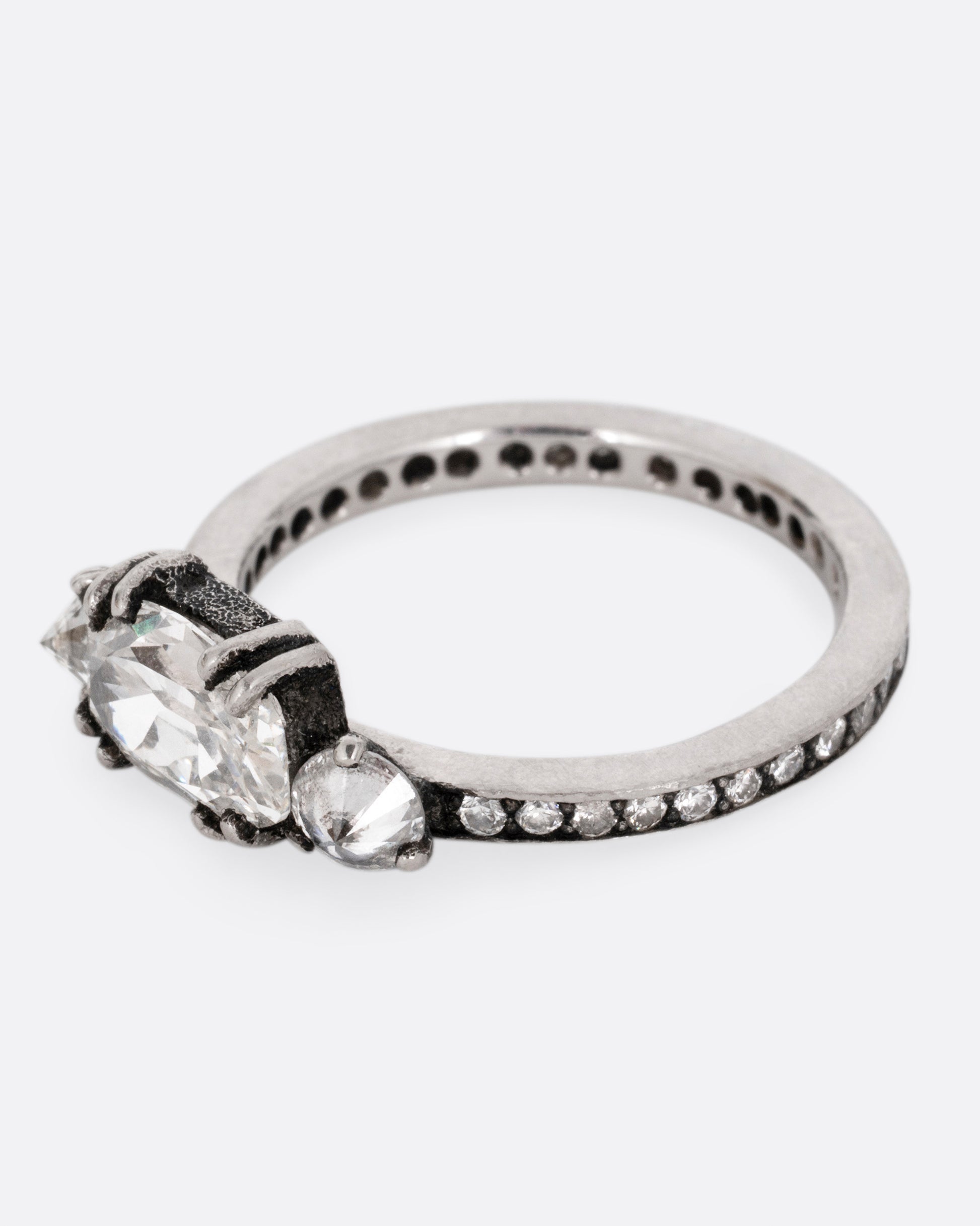 A platinum ring with a trio of diamonds, set upon a contrasting darkened channel of bead set diamonds. The elongated cushion cut center diamond is accented by two reverse set brilliant cut diamonds. Shown from the side. 