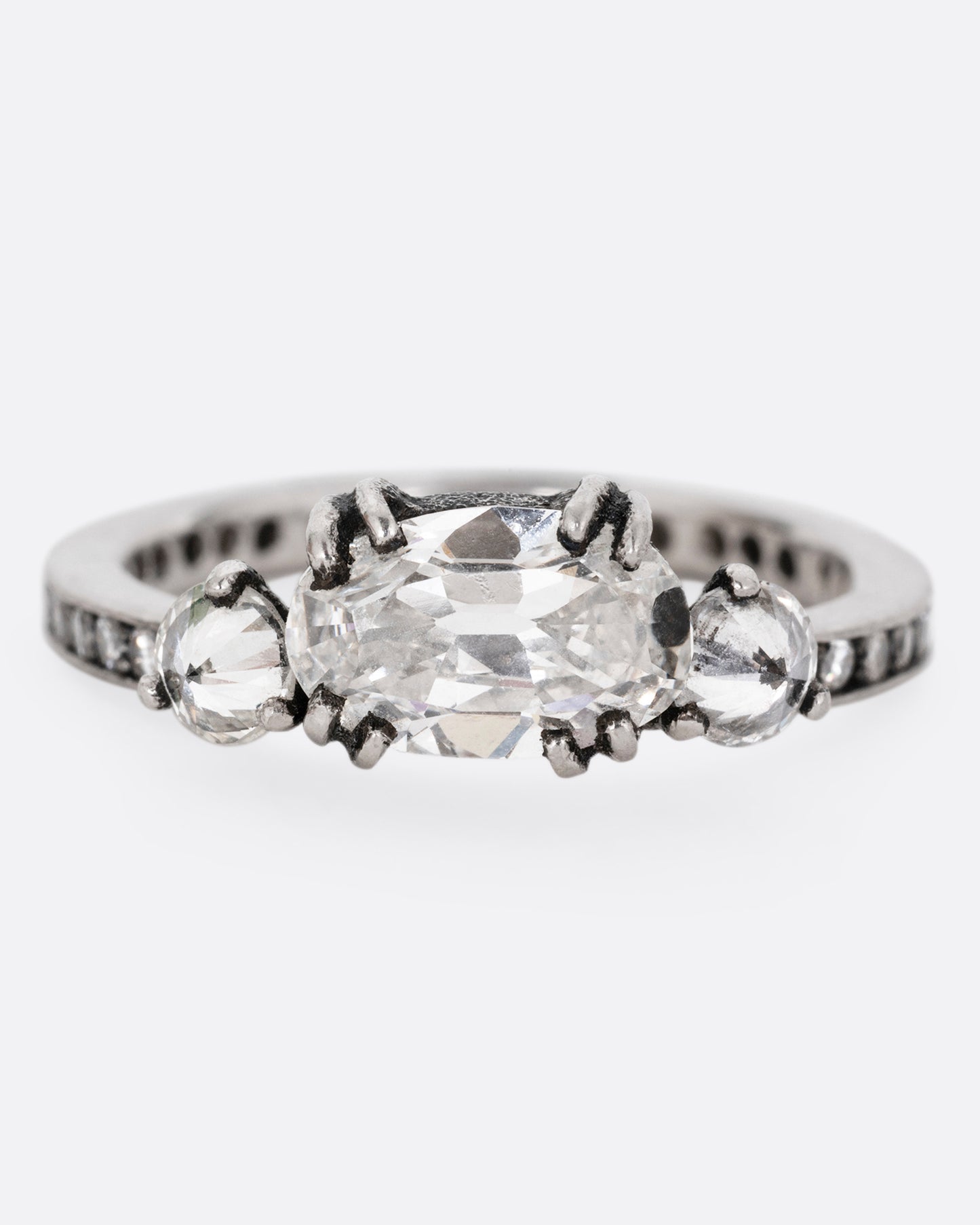 A platinum ring with a trio of diamonds, set upon a contrasting darkened channel of bead set diamonds. The elongated cushion cut center diamond is accented by two reverse set brilliant cut diamonds. Shown from the front. 