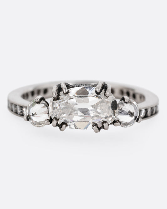 A platinum ring with a trio of diamonds, set upon a contrasting darkened channel of bead set diamonds. The elongated cushion cut center diamond is accented by two reverse set brilliant cut diamonds. Shown from the front. 