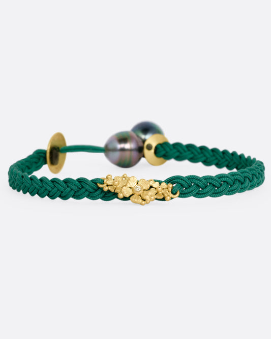 A braided green cord bracelet with a yellow gold cluster of diamond accented flowers and a Tahitian pearl toggle closure. Shown from the front.