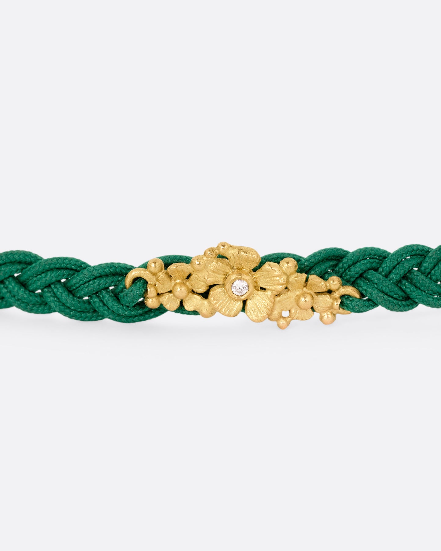 A close up of a braided green cord bracelet with a yellow gold cluster of diamond accented flowers and a Tahitian pearl toggle closure. 