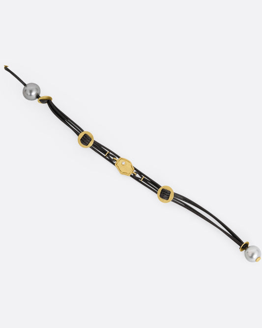 A  black cord bracelet with a diamond studded tag flanked by two gold slides and a pearl closure. Shown open from the front.