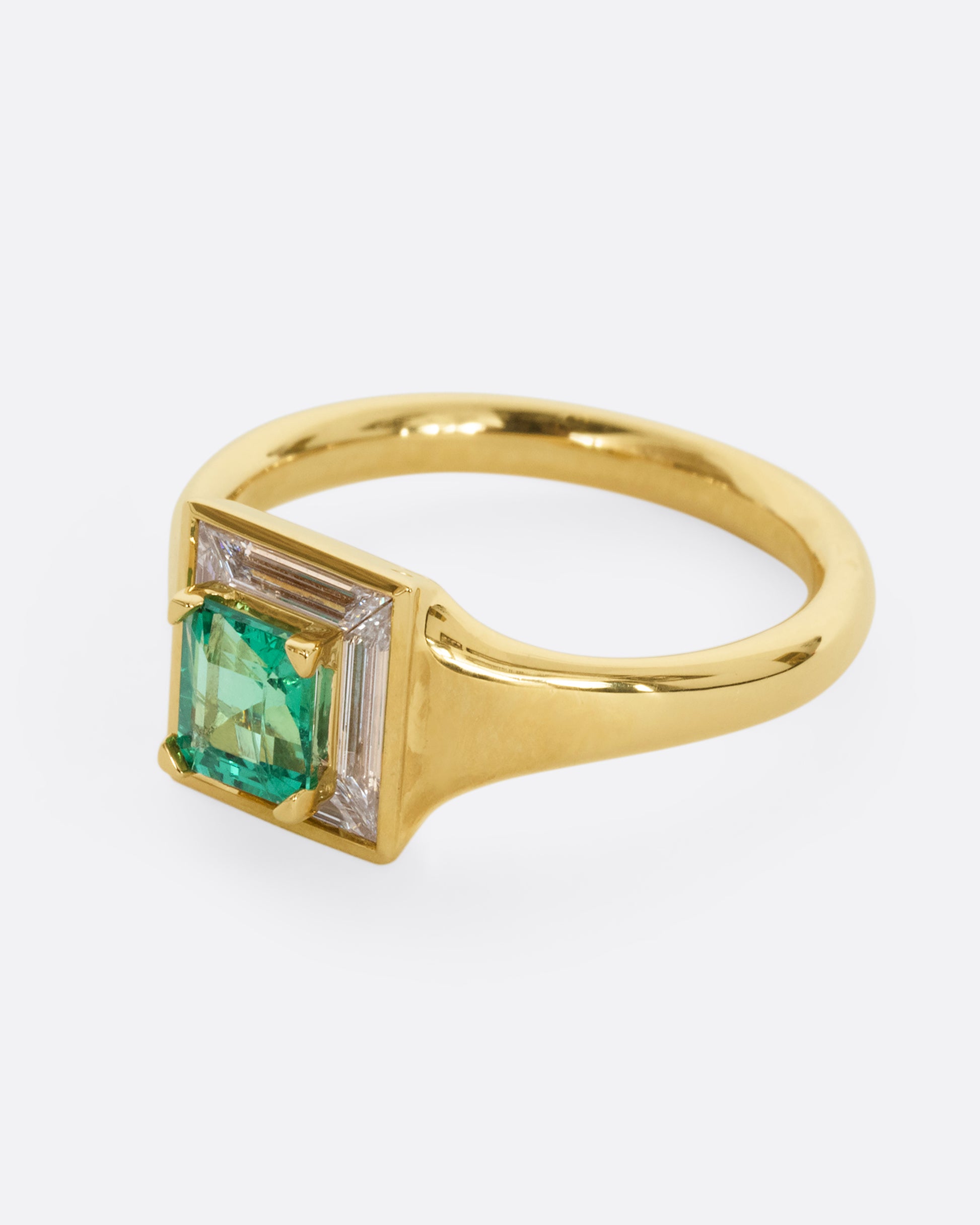 A yellow gold ring with a square emerald at the center surrounded by white baguette diamonds. Shown from the side.