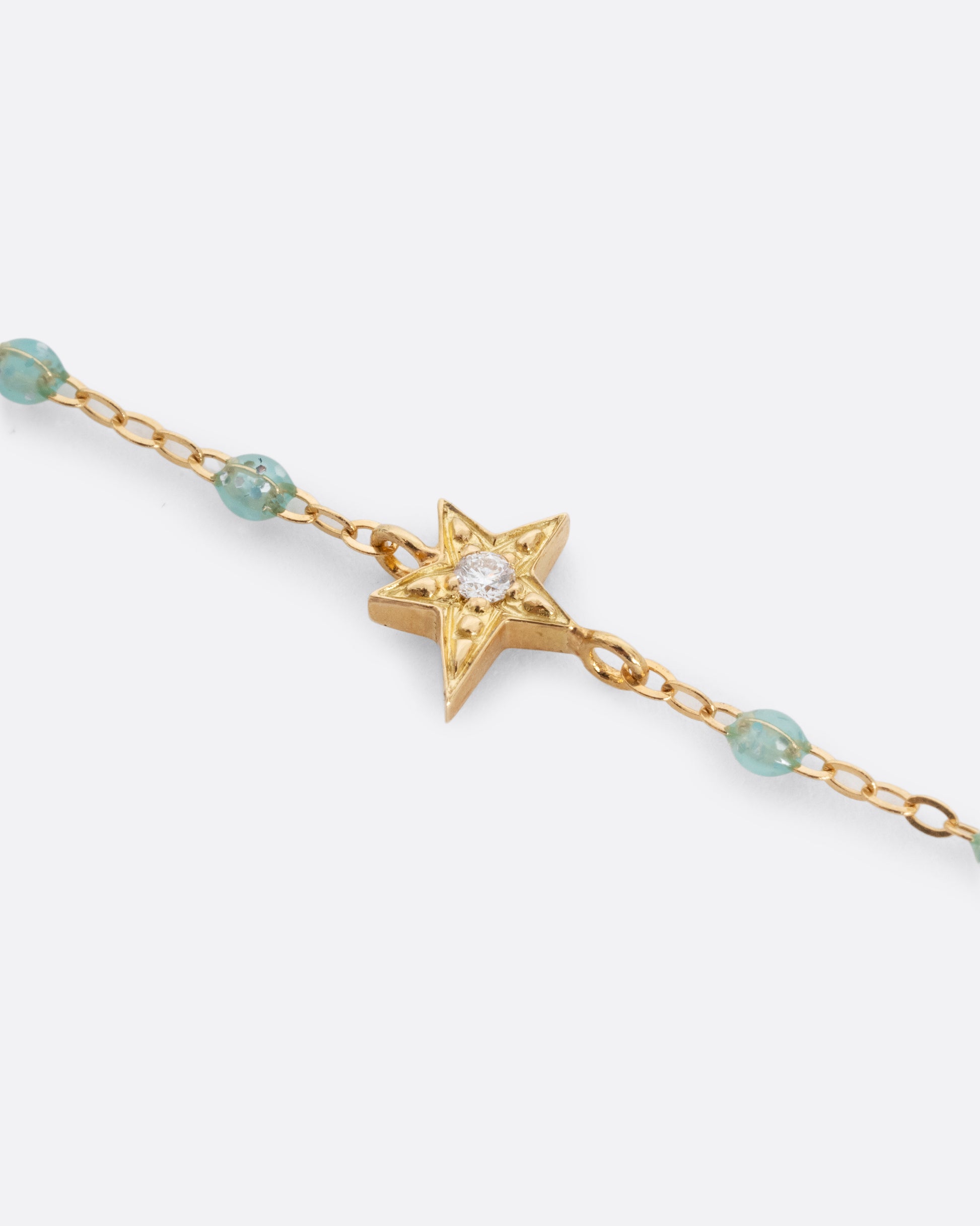 A close up shot of a yellow gold cable chain bracelet with droplets of  aqua blue resin and a yellow gold star charm. A single white diamond is set at the center of the star. Shown from the front.