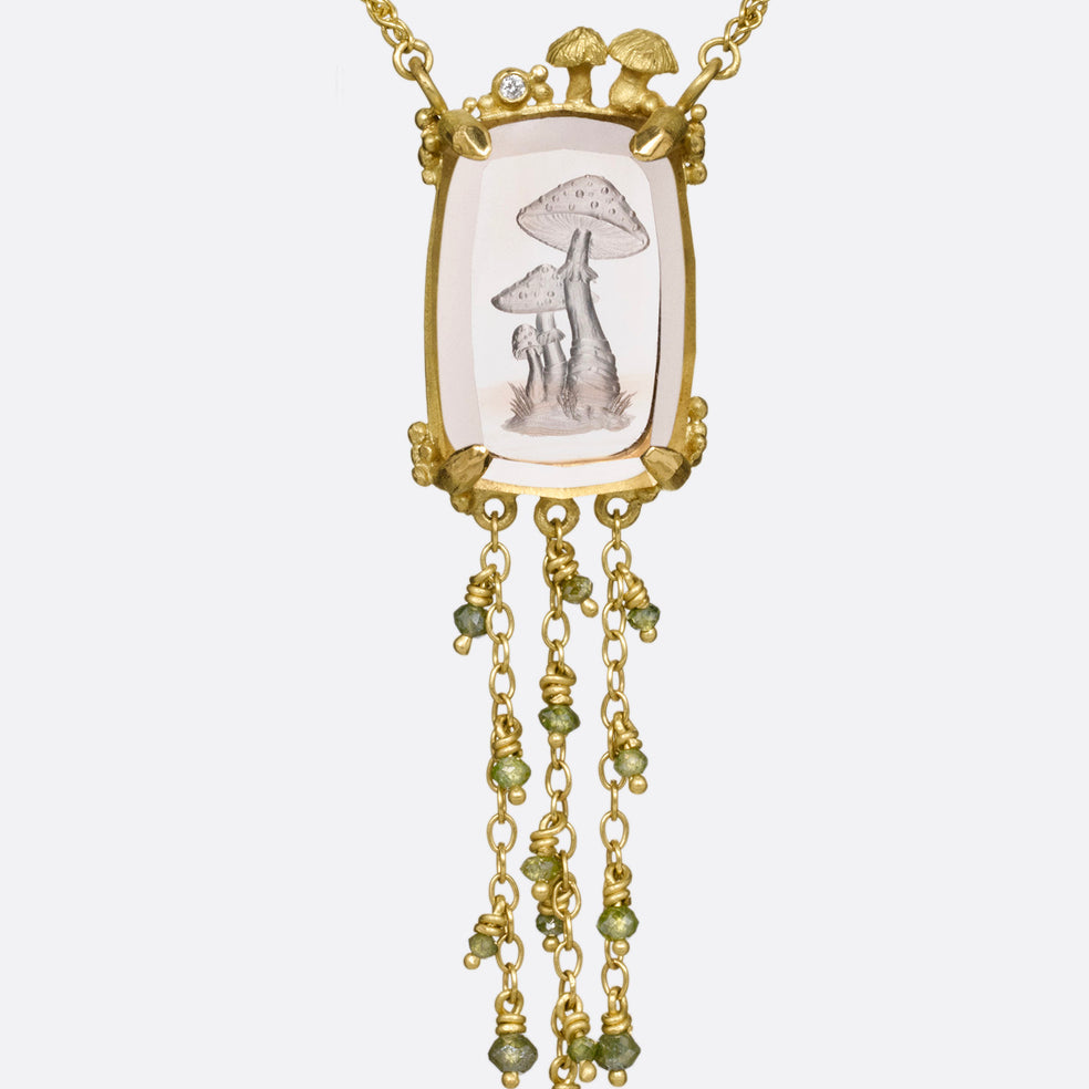 A yellow gold pendant necklace with a hand carved cluster of mushrooms in a light pink topaz with gold mushrooms and a diamond at the top edge. Three gold chains with green diamond beads hand from its base. Shown from the front.