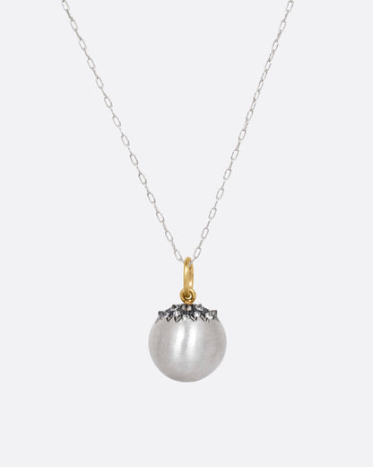 A silver cable chain necklace with a sizable sterling orb pendant peppered with grey reverse set diamonds and a yellow gold bail, shown from the front.