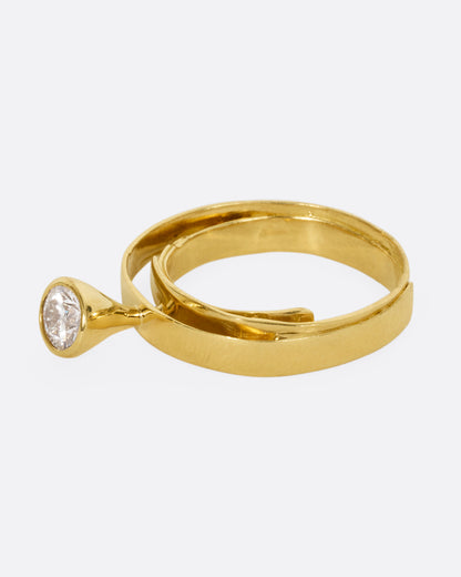A yellow gold ring that loops over itself twice with a tall bezel set diamond at its center. Shown from the side.