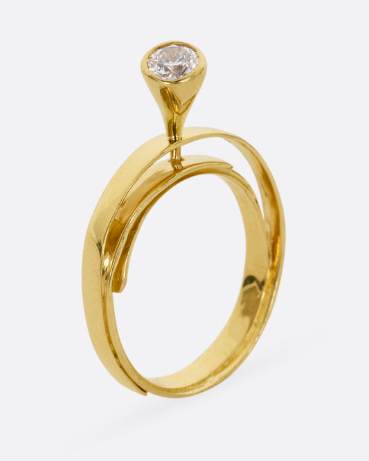 A yellow gold ring that loops over itself twice with a tall bezel set diamond at its center. Shown from the side.