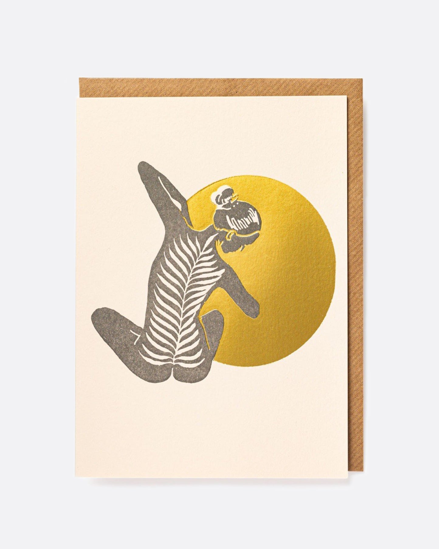 A pale pink greeting card with the silhouette of a person doing yoga in front of a metallic gold orb on the front, the person's spine made from a palm leaf. The card is blank inside.