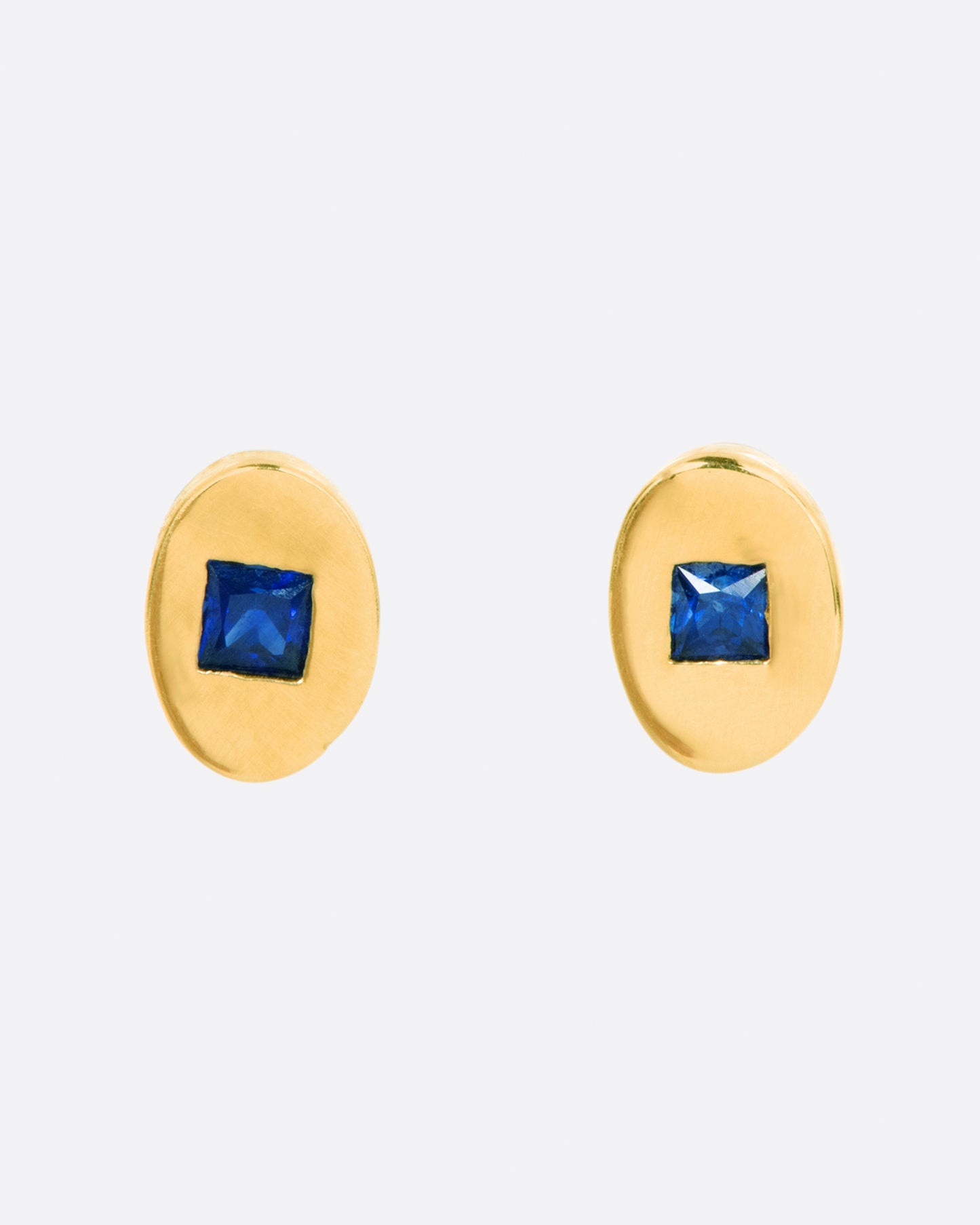 A pair of oval yellow gold stud earrings, each with a princess cut blue sapphire at its center.