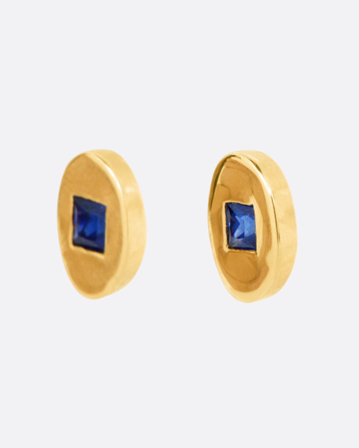 A pair of oval yellow gold stud earrings, each with a princess cut blue sapphire at its center. Shown from the side.