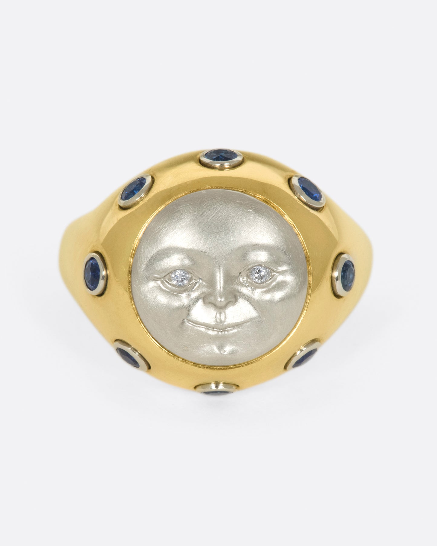 A platinum moonface sunken into a gold ring, surrounded by sapphire cabochons.