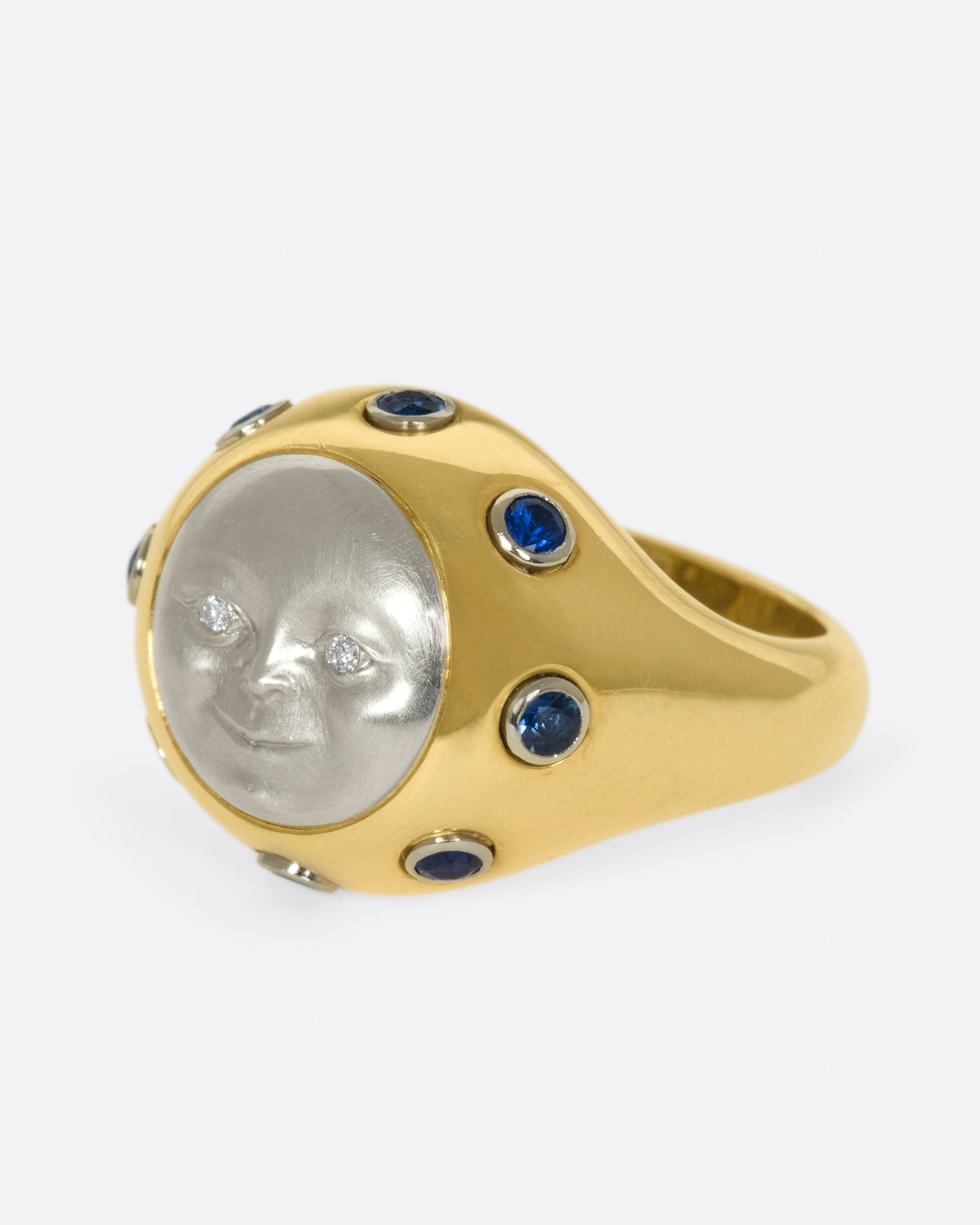 A platinum moonface sunken into a gold ring, surrounded by sapphire cabochons.