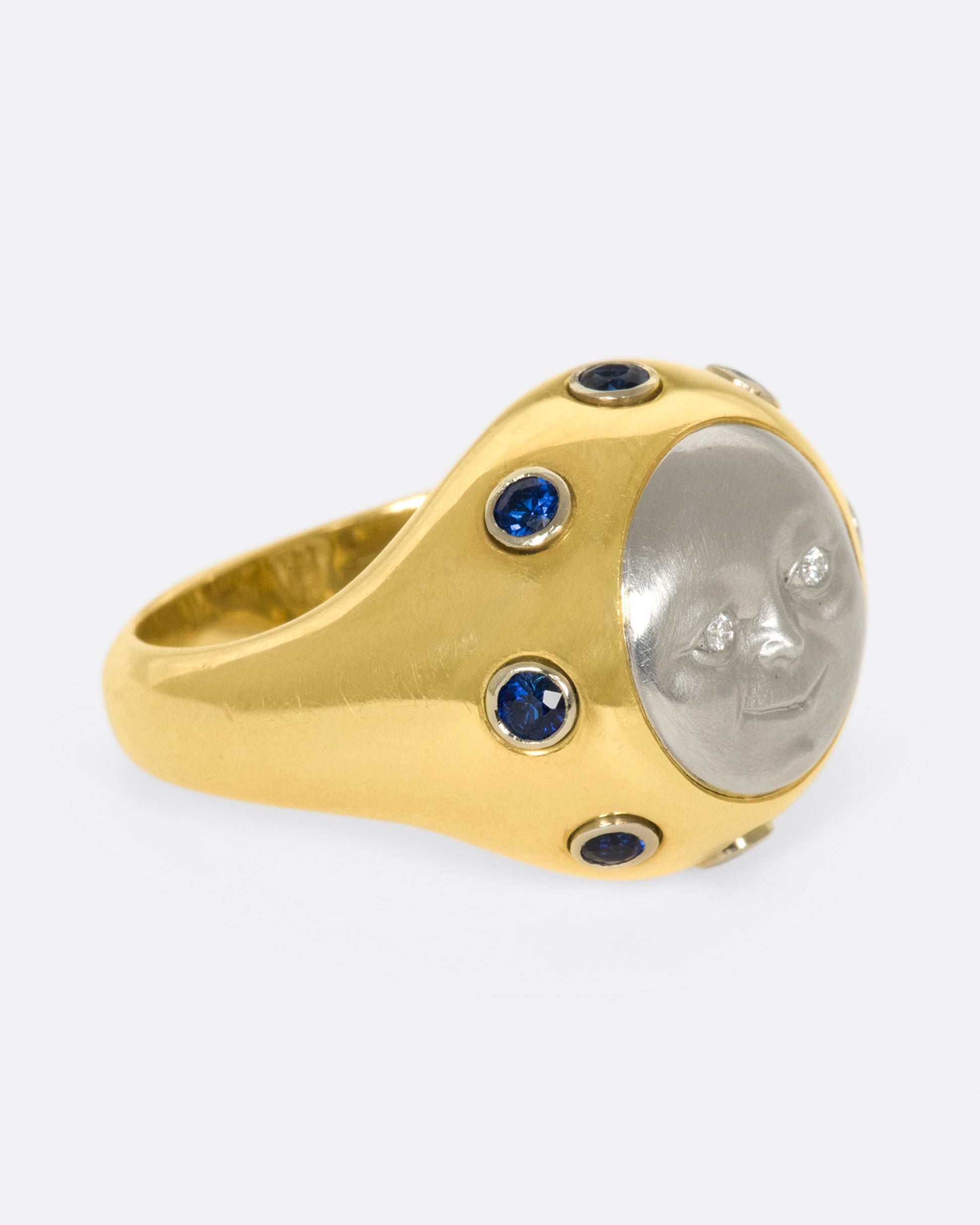 A platinum moonface sunken into a gold ring, surrounded by sapphire cabochons.