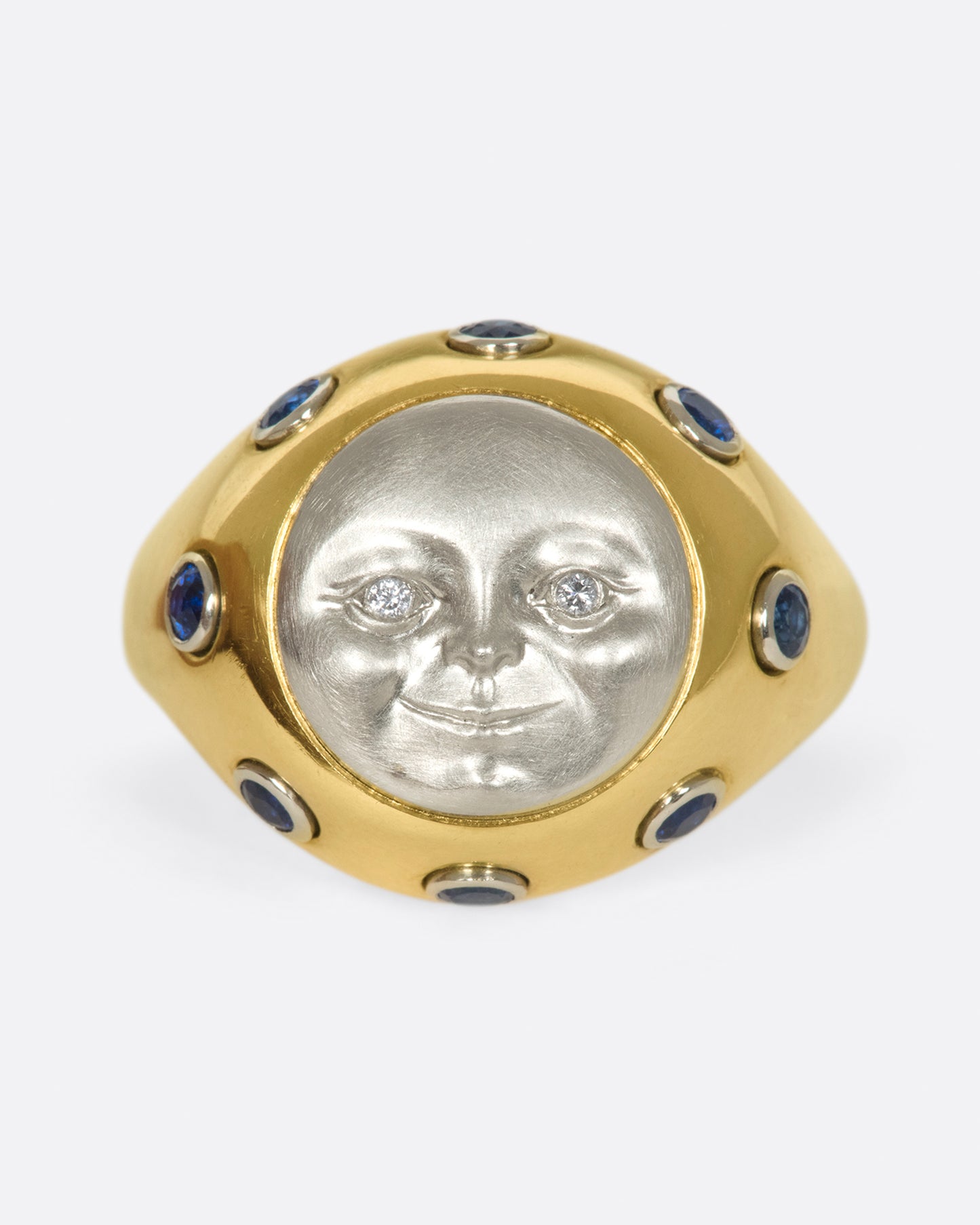 A platinum moonface sunken into a gold ring, surrounded by sapphire cabochons.