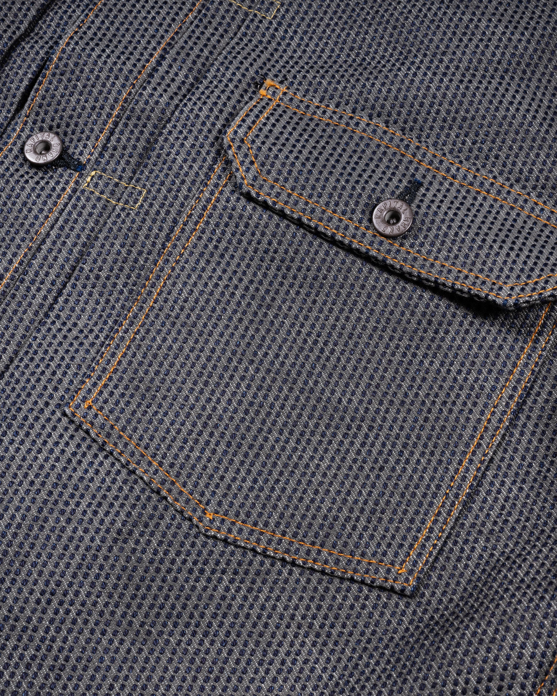 A dark blue textured button down denim jacket with a single pocket. Close up of the pocket.