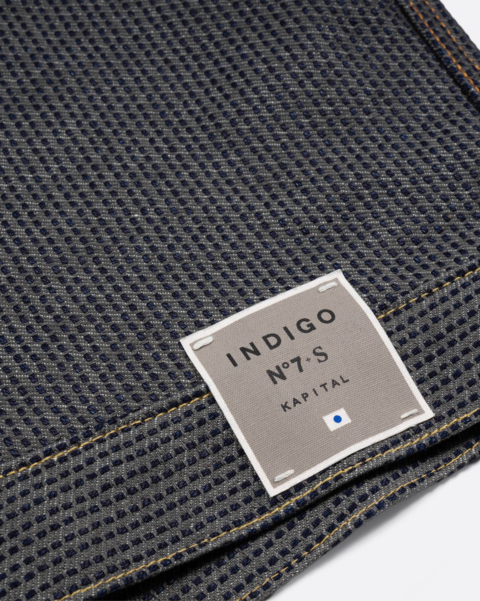 A dark blue textured button down denim jacket with a single pocket. Close up of the label on the hem.