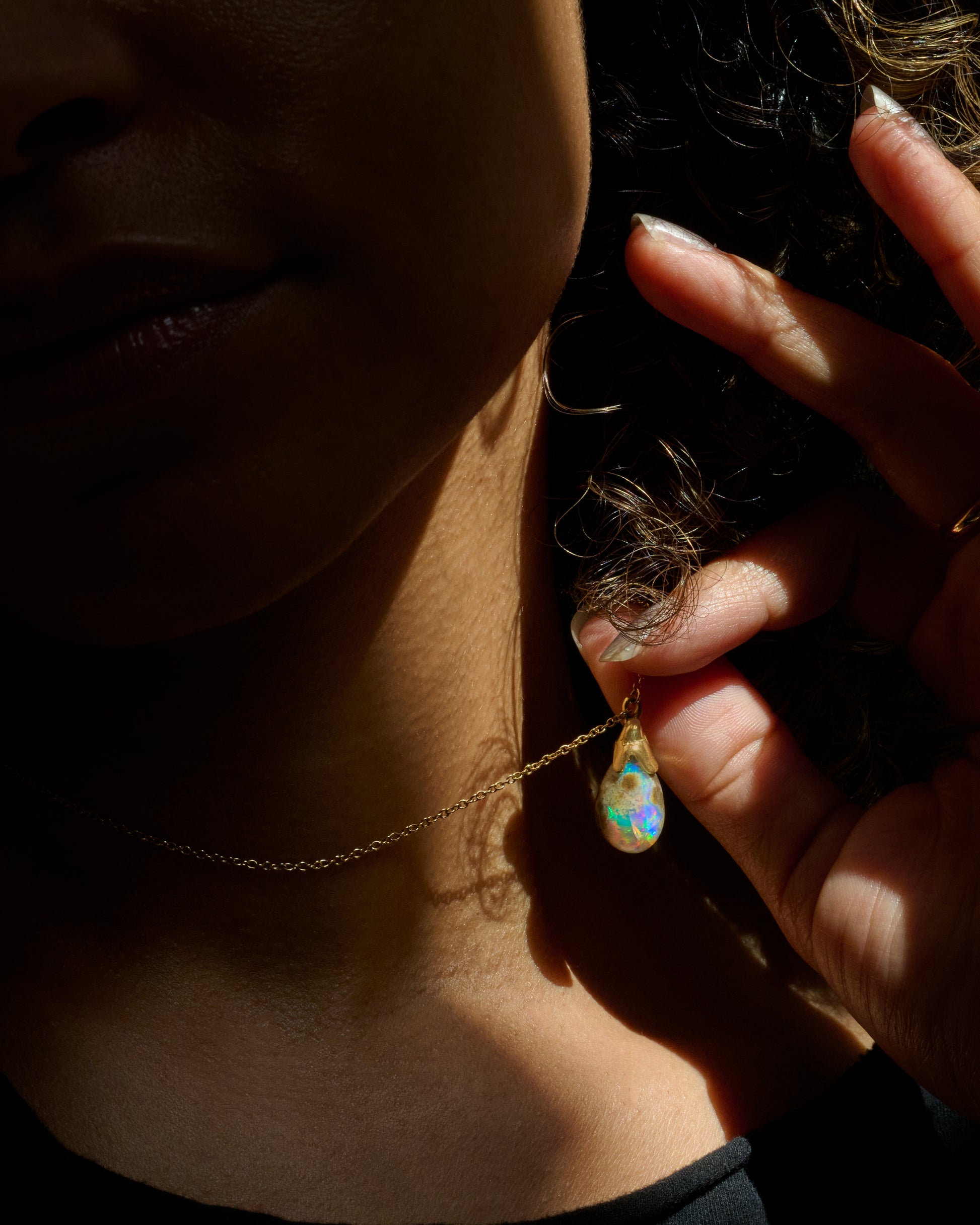 A close-up lifestyle shot of a person wearing a delicate gold chain necklace with a teardrop-shaped iridescent opal pendant, held by a hand in dramatic, high-contrast sunlight.