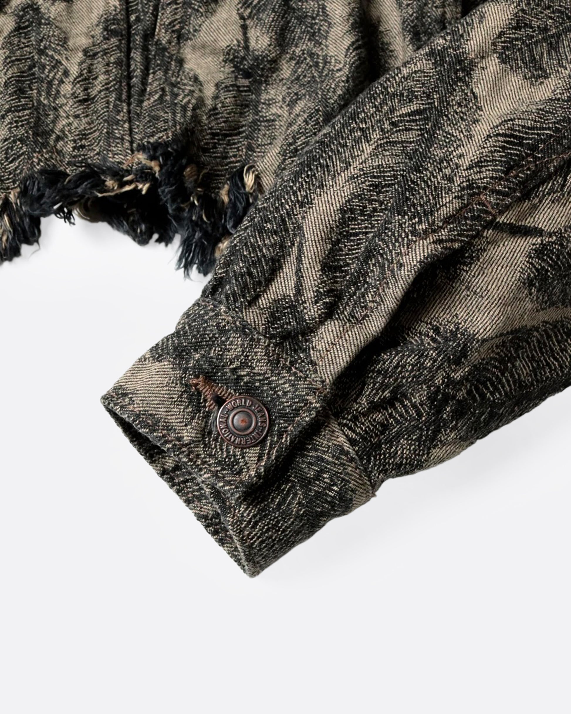 A close up of the cuff of a black and beige jacquard denim jacket with an all-over 3D feather motif and a cut-off hemline and raw feathered seam. 