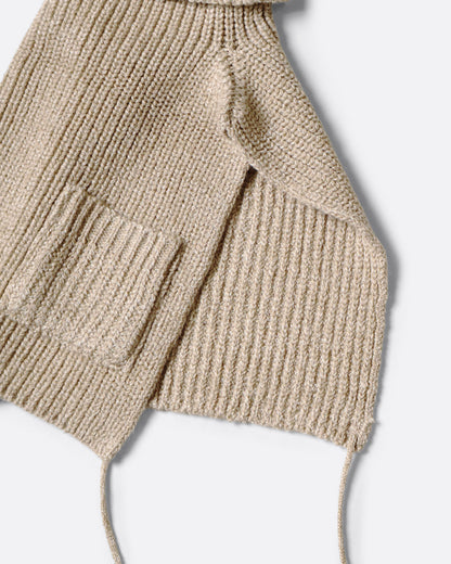 A close up of a beige loose knit bib poncho with a front pocket, long tassels, and a high neck.