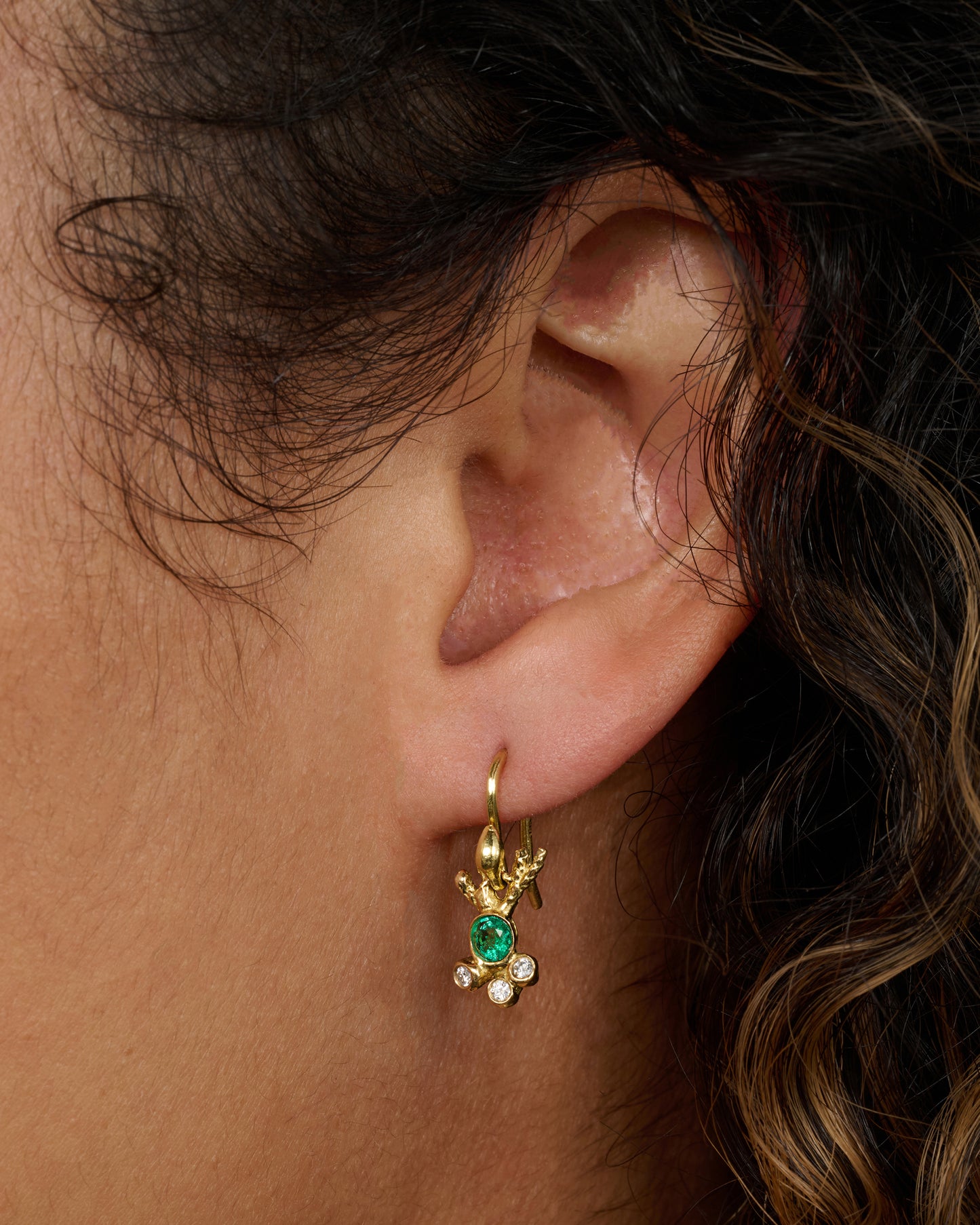 Yellow gold abstract ray earrings with an emerald at the center and three diamonds clustered below. View of a single earring worn.