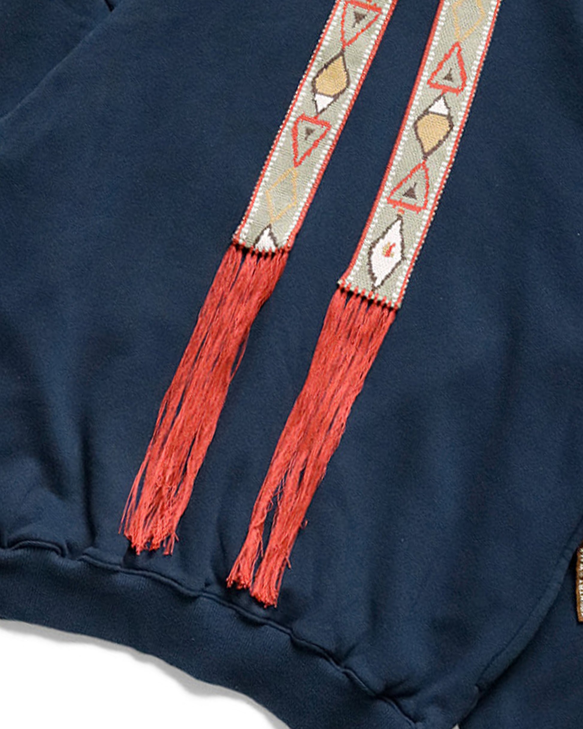 A blue cotton fleece hoodie with an oversized silhouette, accented with wide, fixed drawstrings in a geometric pattern with long red fringe.