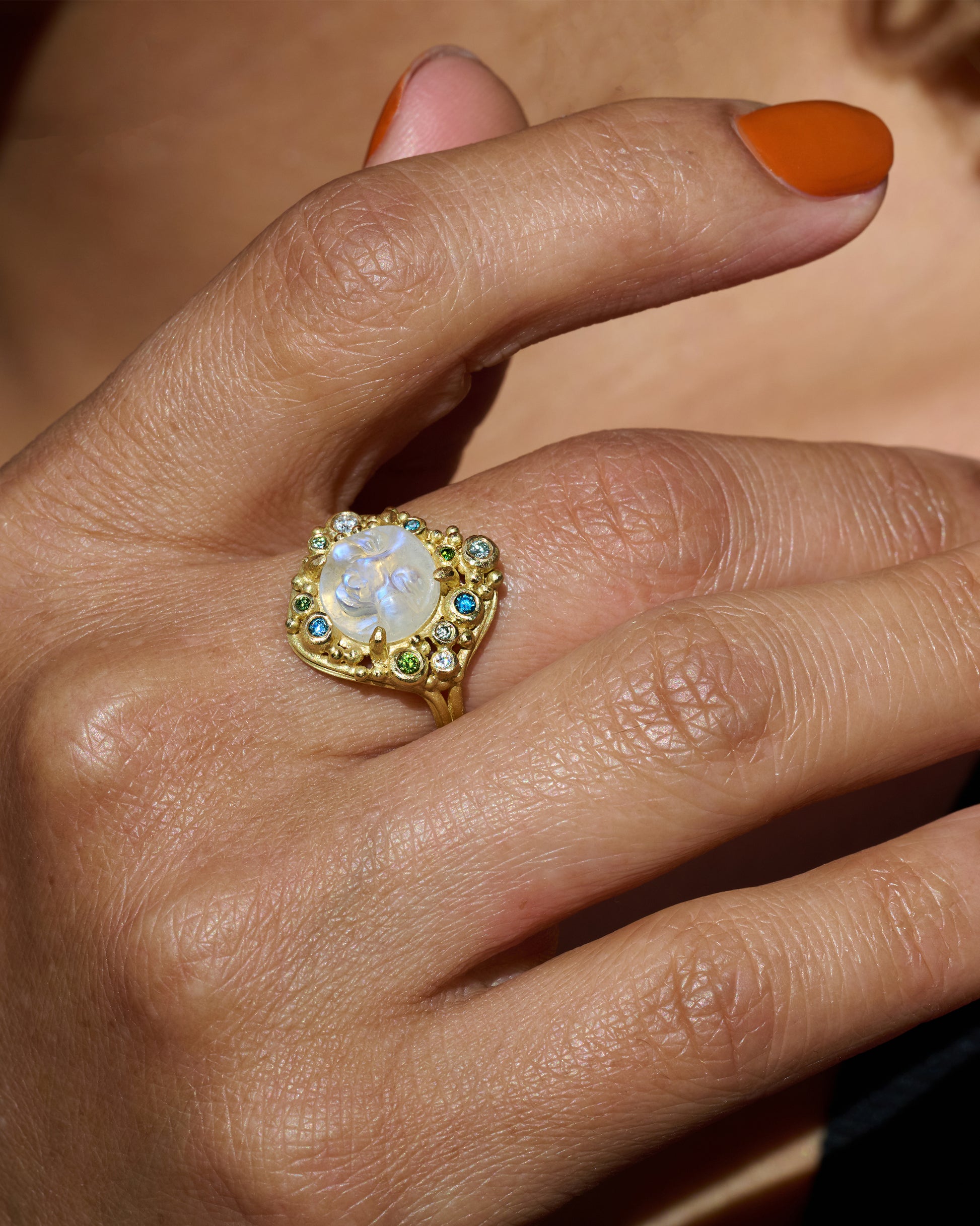 A yellow gold ring featuring a carved moonstone face with a halo of multicolored diamonds. View on a hand.