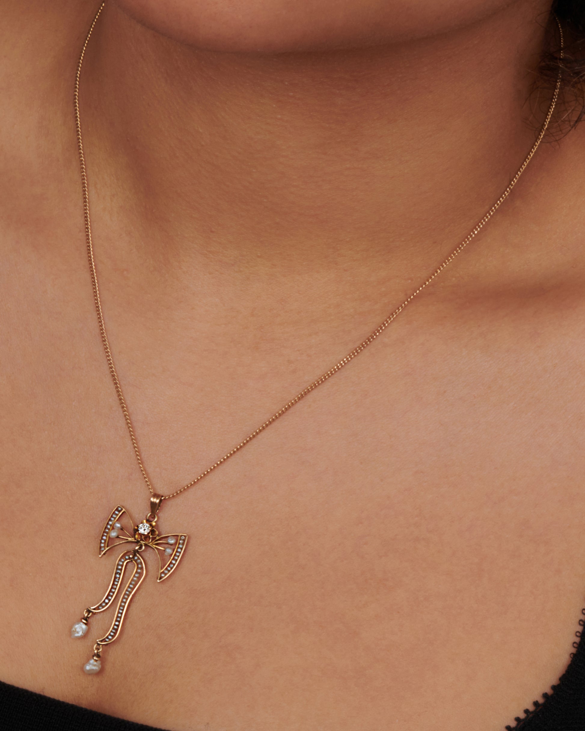 A close up view of a the top half of a yellow gold bow pendant with seed pearls and a diamond. View on neck.