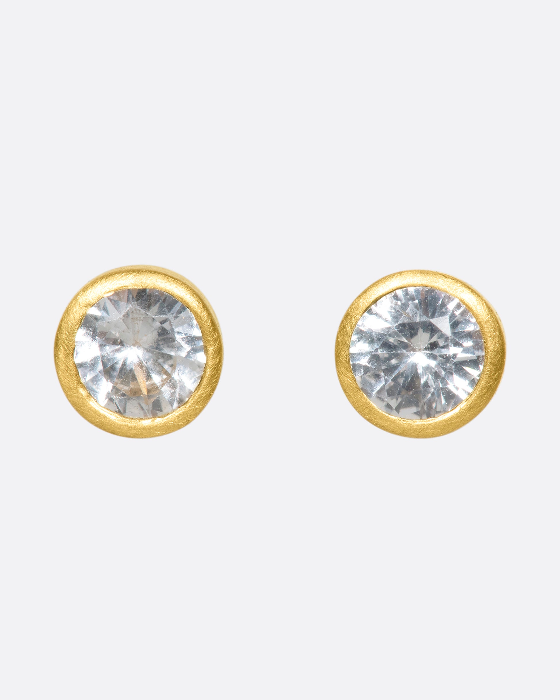 A pair of yellow gold solitaire earrings with round white sapphires in bezel settings.