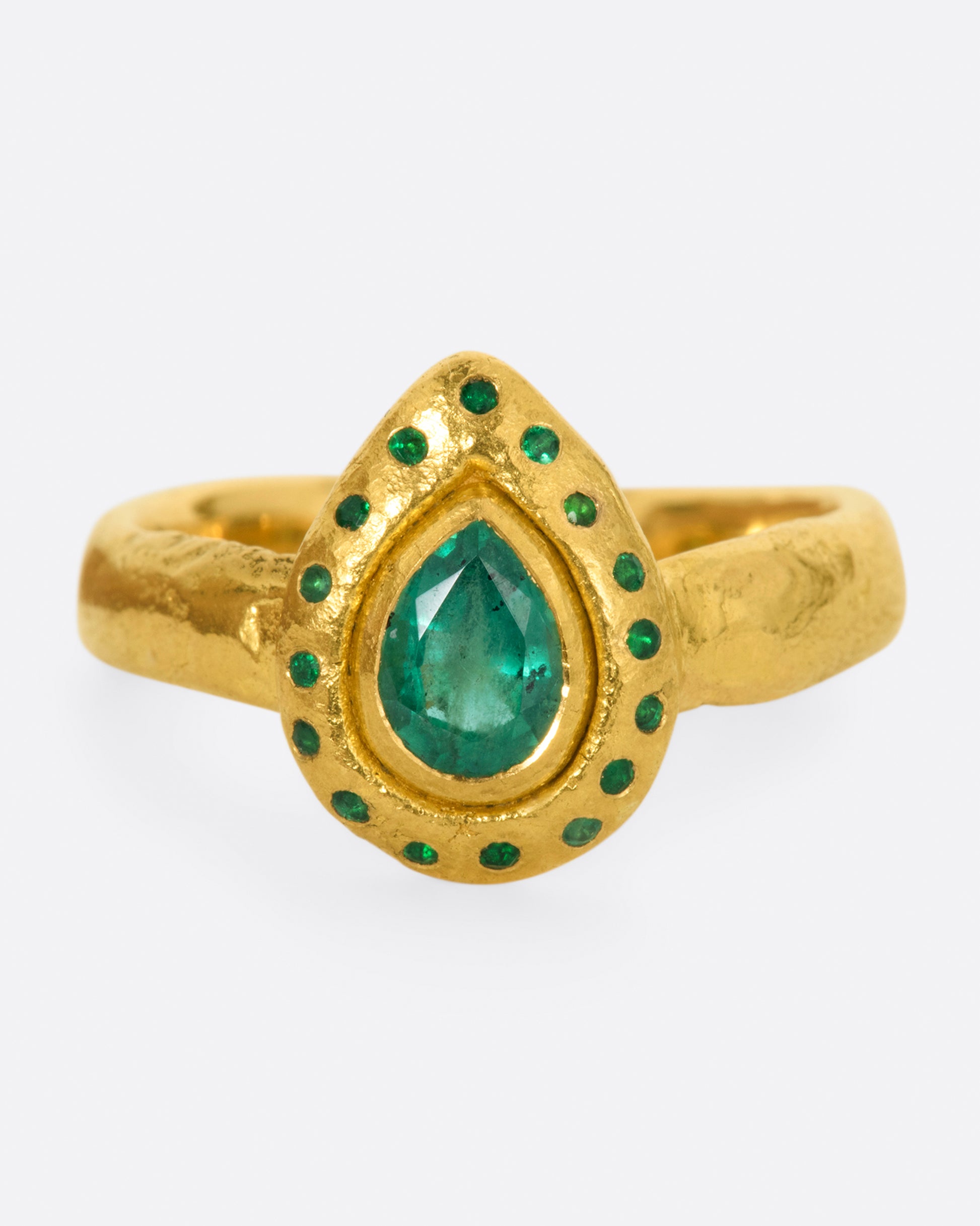 A high karat gold ring with an organic shaped band and a pear shaped emerald at its center with a sunken round emerald halo.