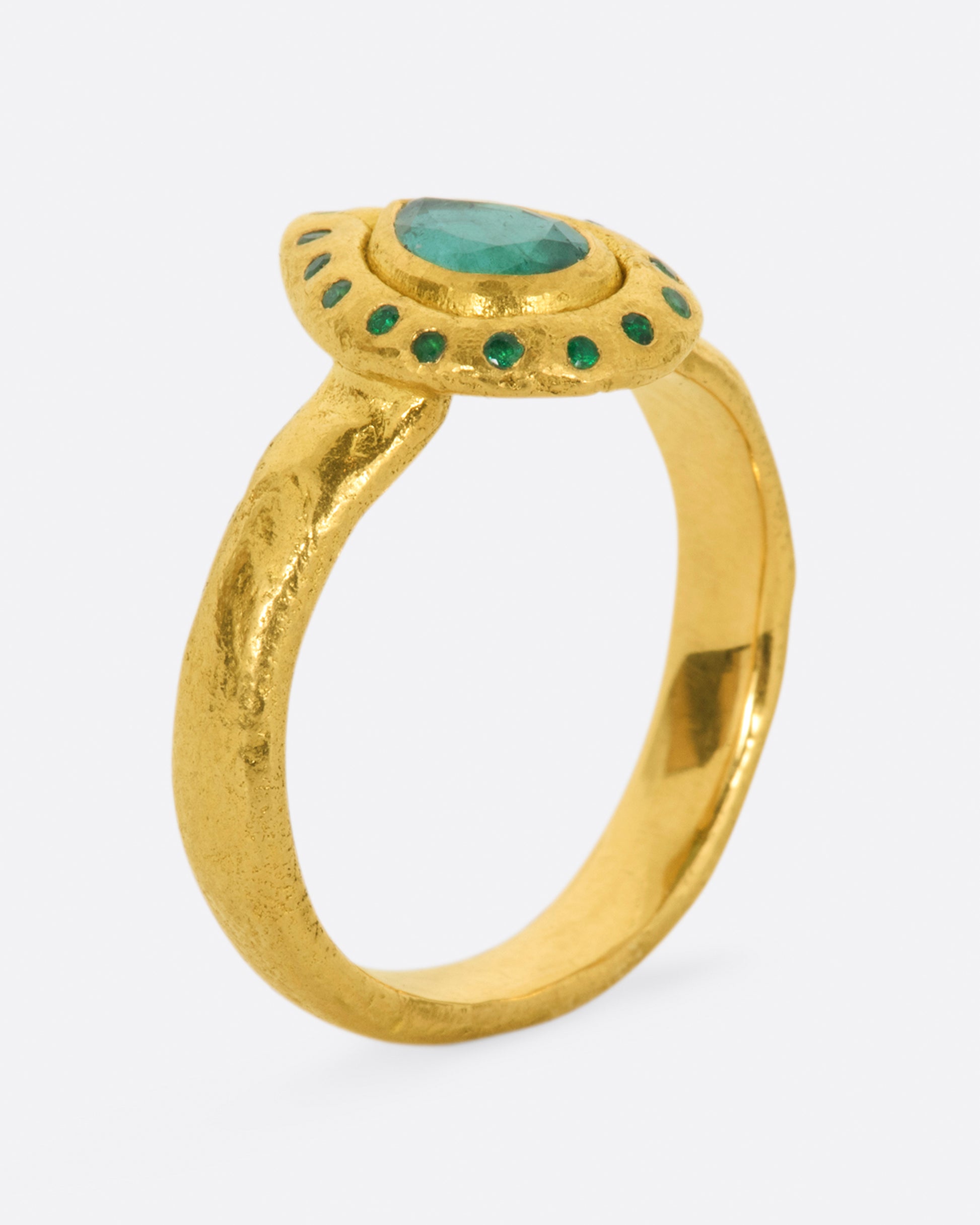 A high karat gold ring with an organic shaped band and a pear shaped emerald at its center with a sunken round emerald halo. Shown from the side.