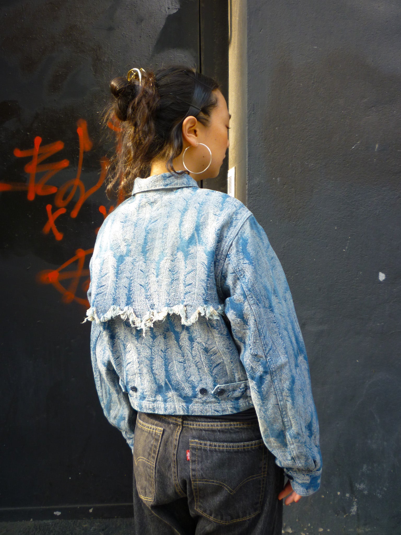 A light blue jacquard denim jacket with  an all- over feather motif, and a cut-off hemline with raw feathered seam. Shown from the back on a person.