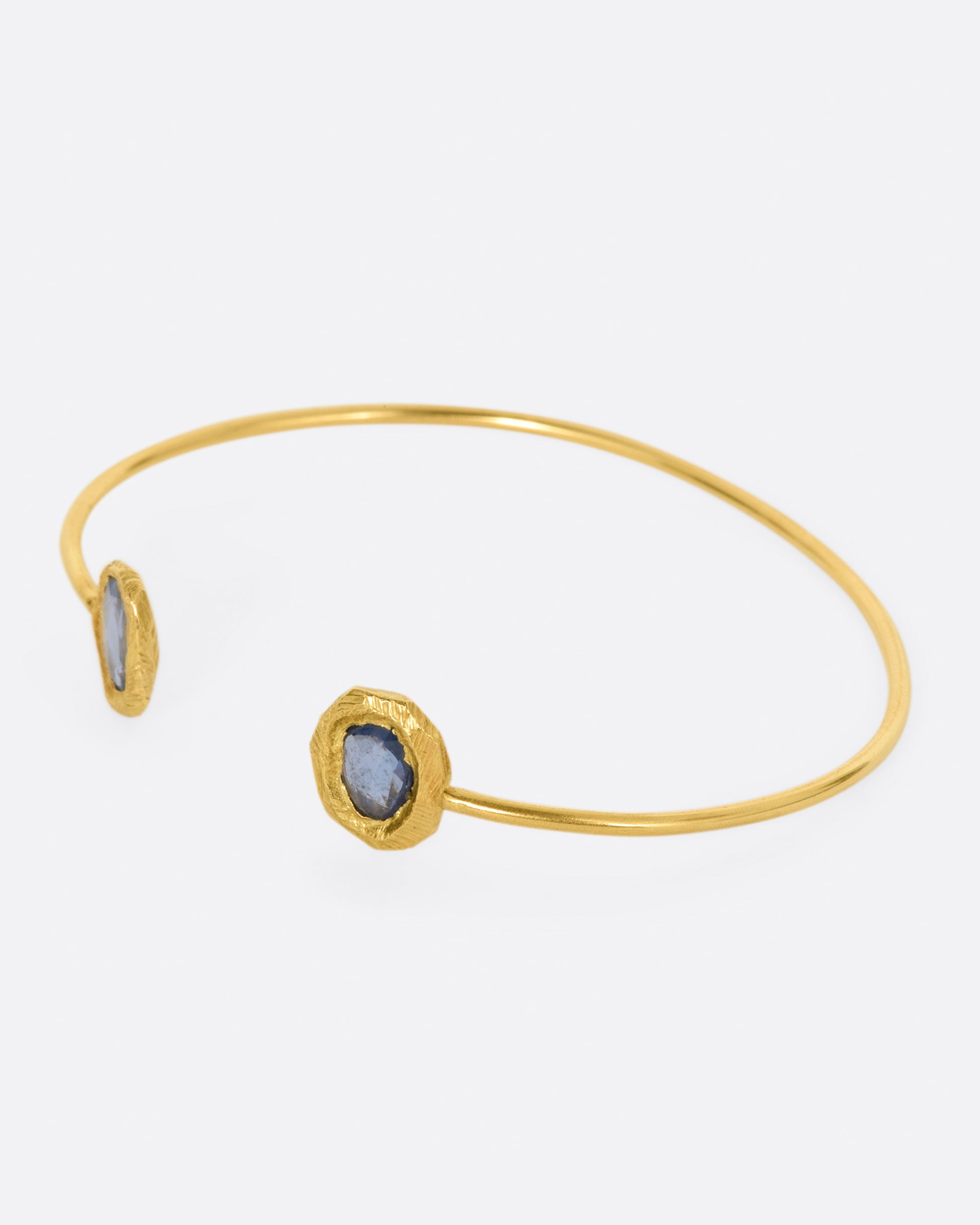 A simple yellow gold wire cuff bracelet with a blue sapphire in a hand carved bezel on either end. Shown from the side.