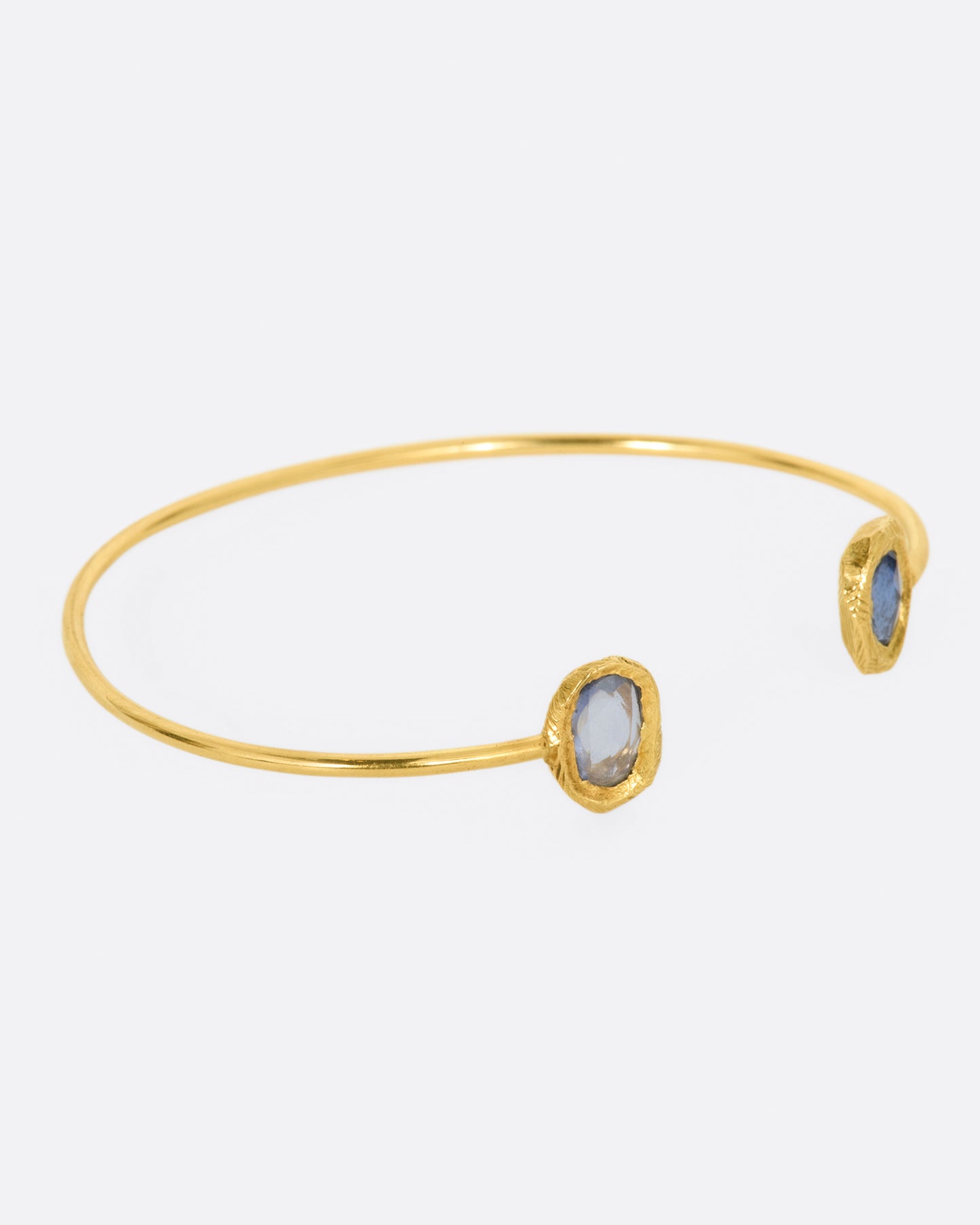 A simple yellow gold wire cuff bracelet with a blue sapphire in a hand carved bezel on either end. Shown from the side.