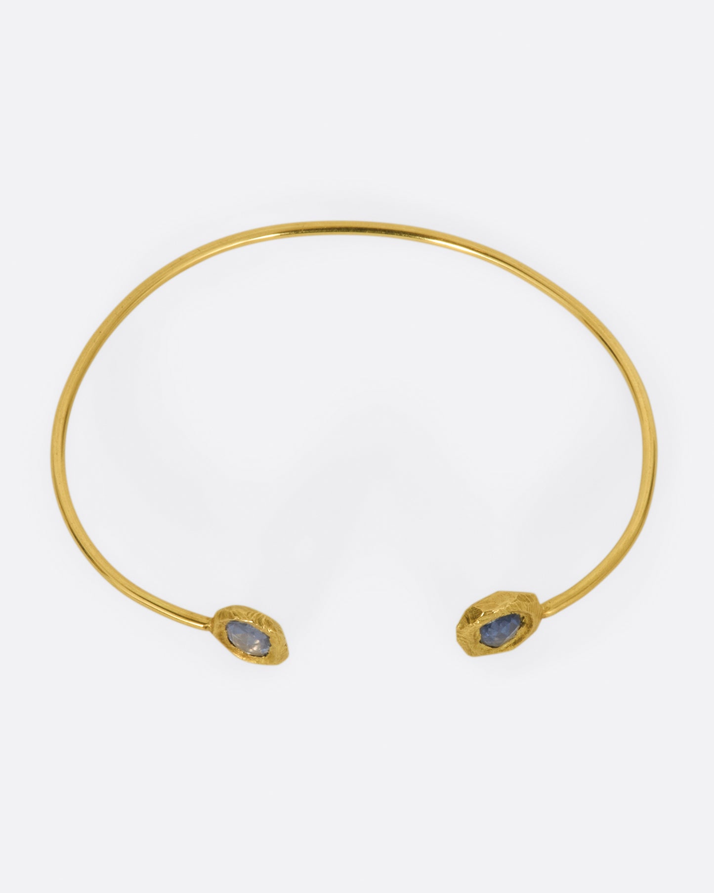 A simple yellow gold wire cuff bracelet with a blue sapphire in a hand carved bezel on either end. Shown from above.