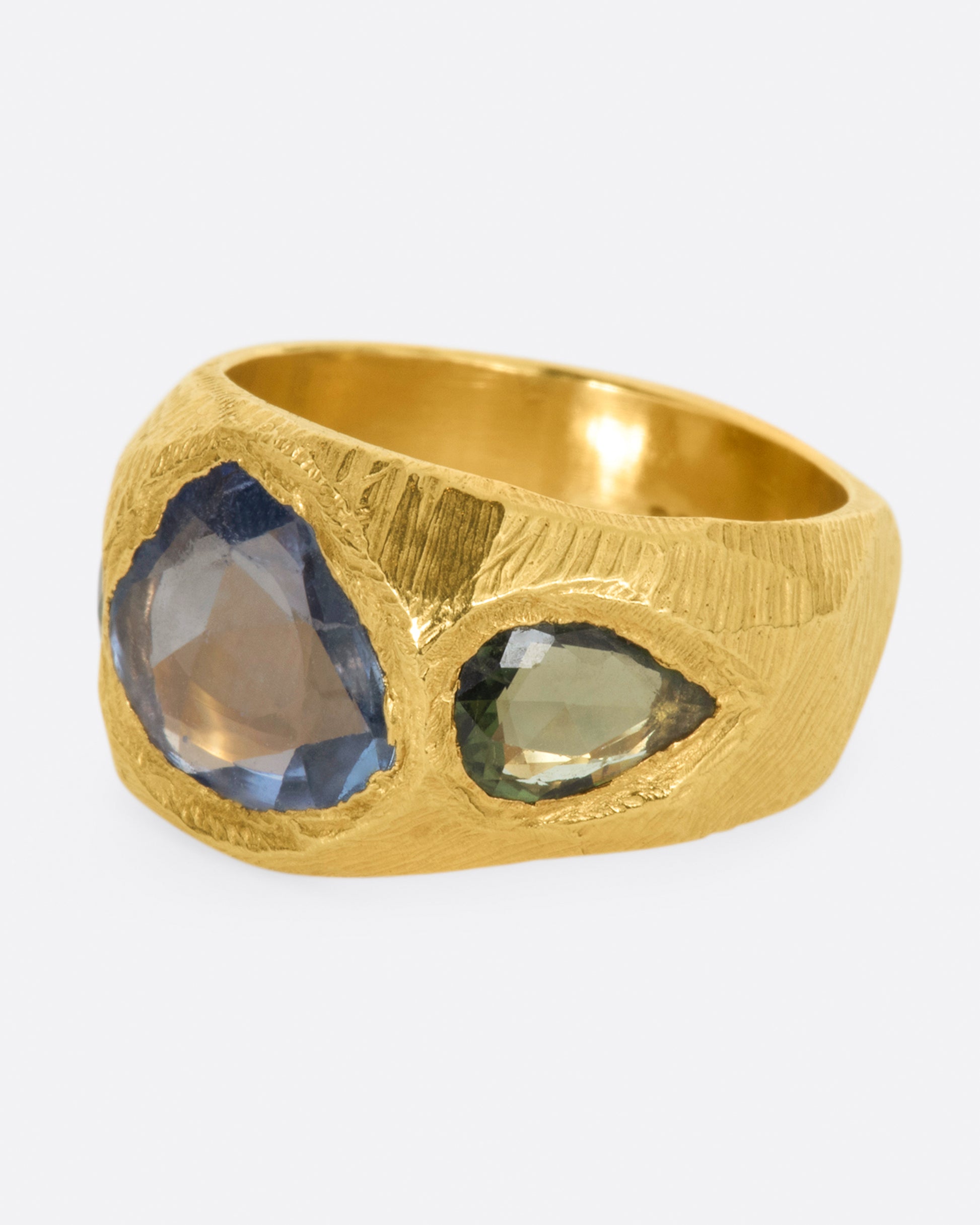 A brushed texture yellow gold ring with a large rose cut blue sapphire at its center with one green pear shaped sapphire on either side. Shown from the side.