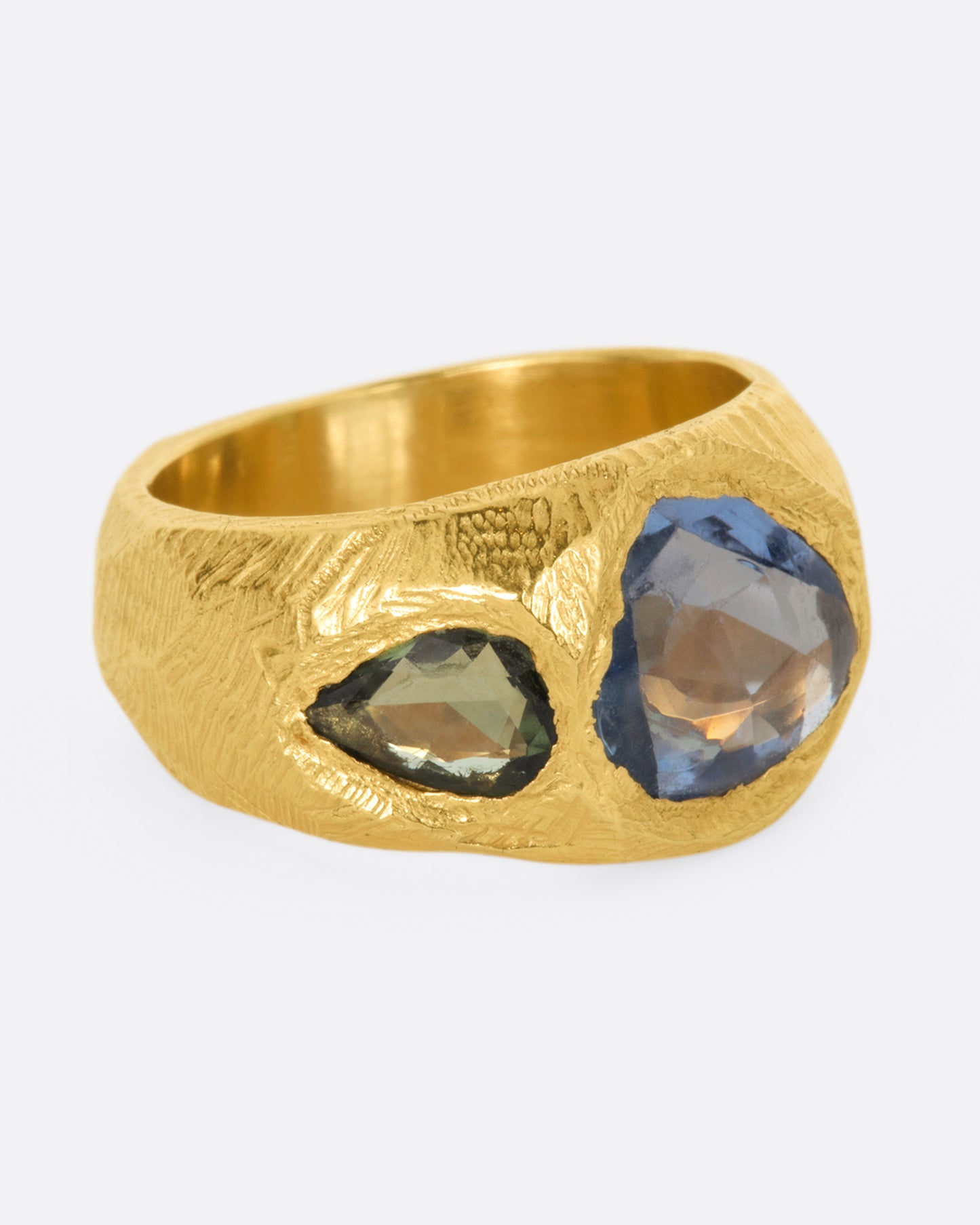 A brushed texture yellow gold ring with a large rose cut blue sapphire at its center with one green pear shaped sapphire on either side. Shown from the side.