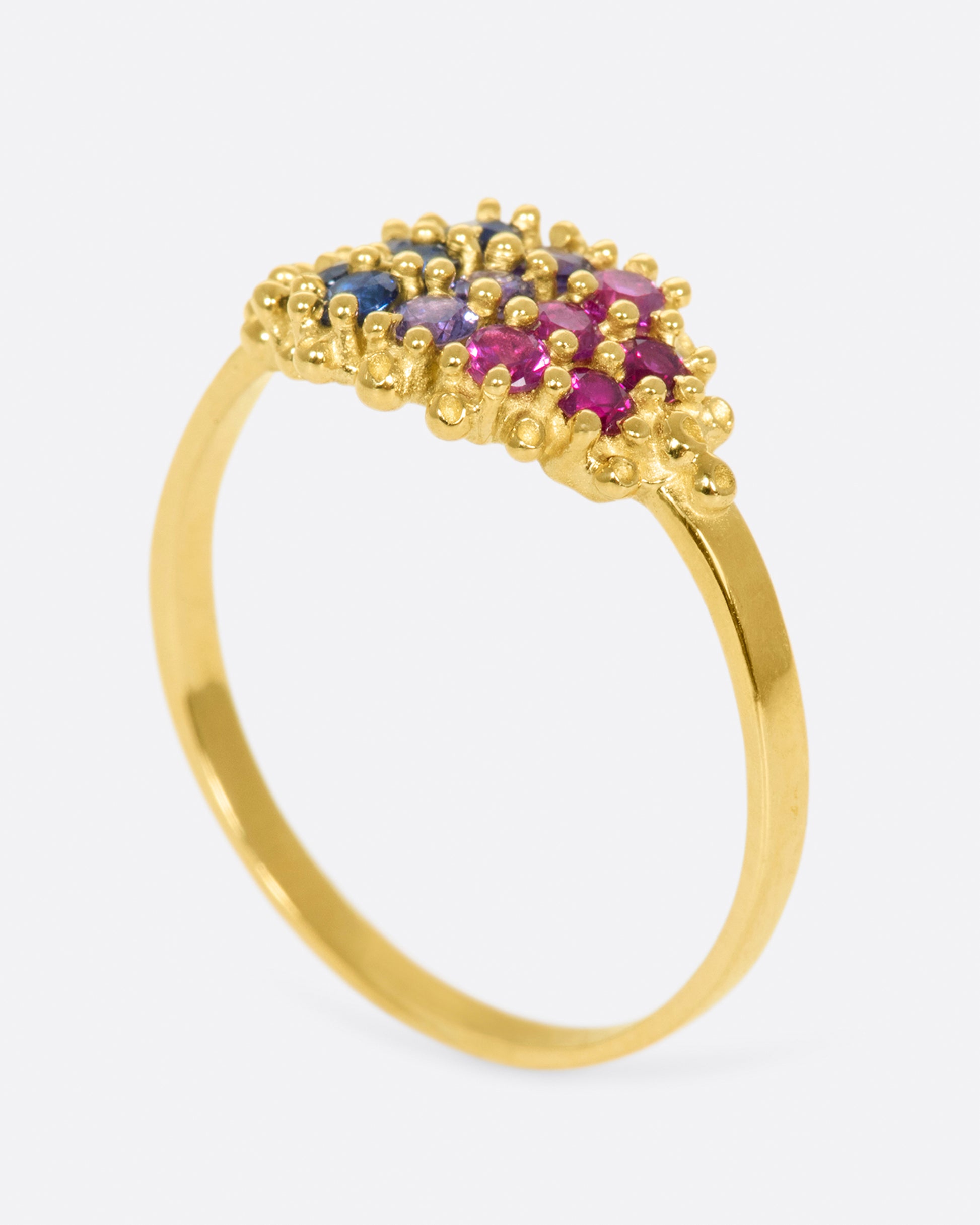 A yellow gold signet style ring whose face is lined with ombré pink to blue sapphires in textured prong settings. Shown from the side.