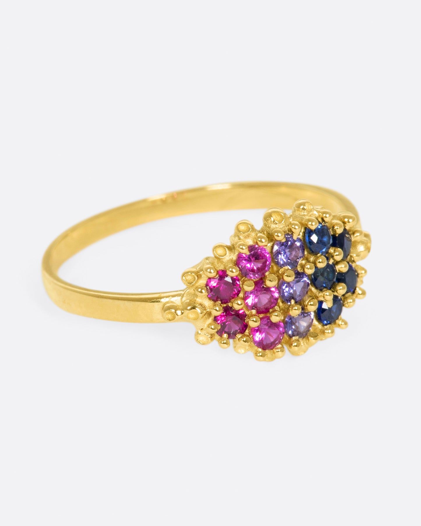 A yellow gold signet style ring whose face is lined with ombré pink to blue sapphires in textured prong settings. Shown from the side.