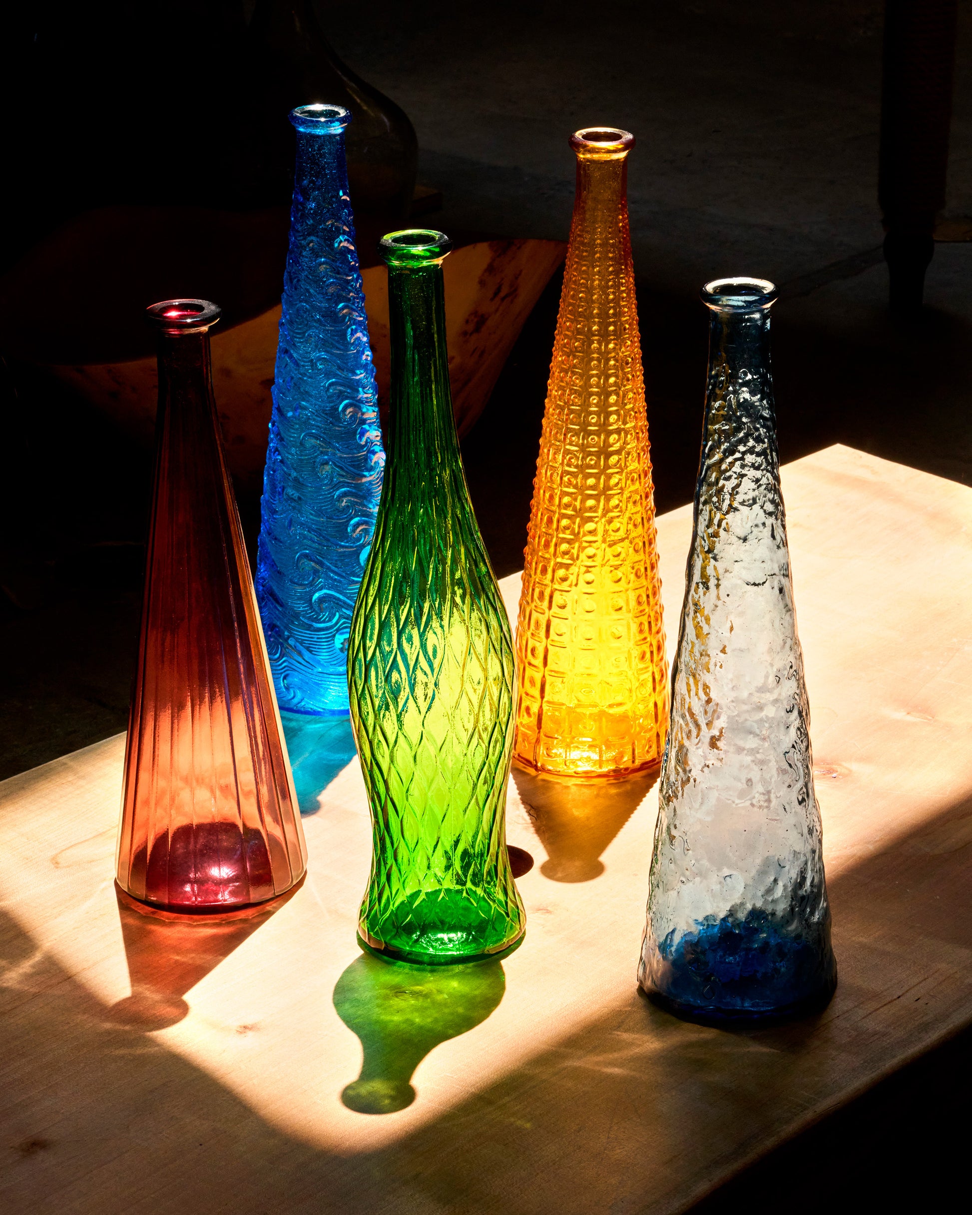 Five tall colorful glass vases, shown from the front in a sunny patch of light.