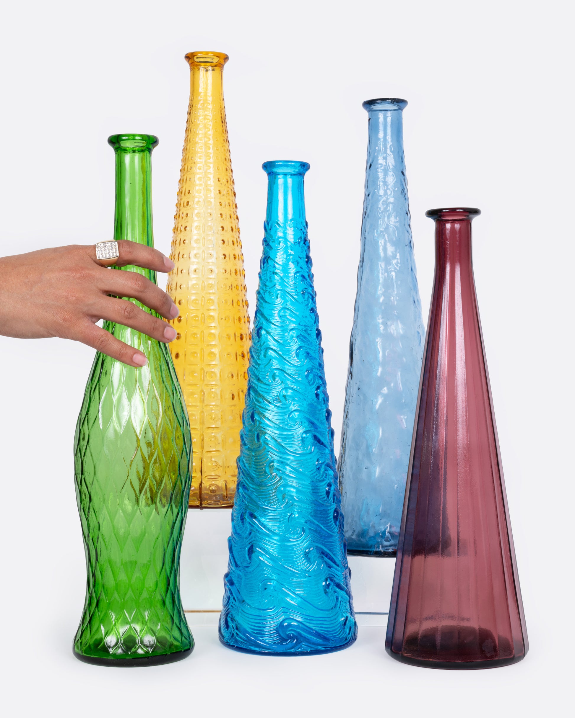 A grouping of five colorful tall glass vases shown from the front with a hand grabbing one.