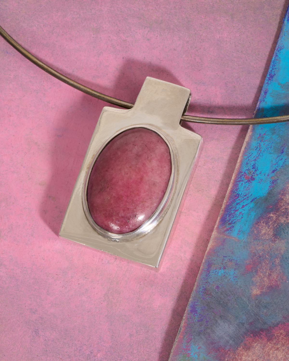 A sterling silver rectangular pendant with an oval pink stone cabochon on a silver wire collar necklace laying flat on a painted pink and blue background.
